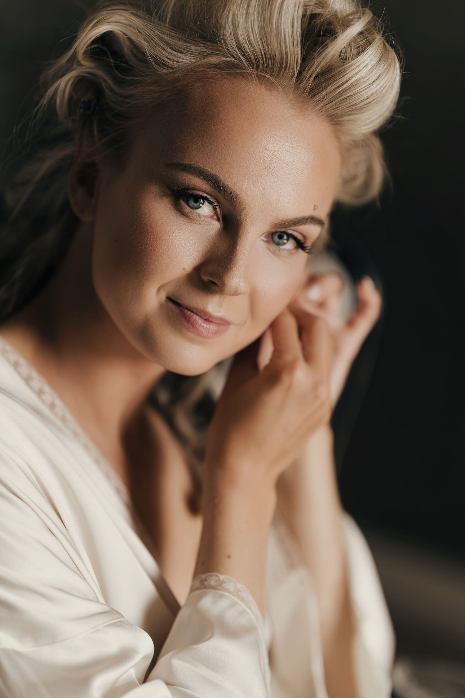 Smiling blonde woman with an elegant updo and a cream satin robe gently touching her face in a soft, warm-toned portrait.