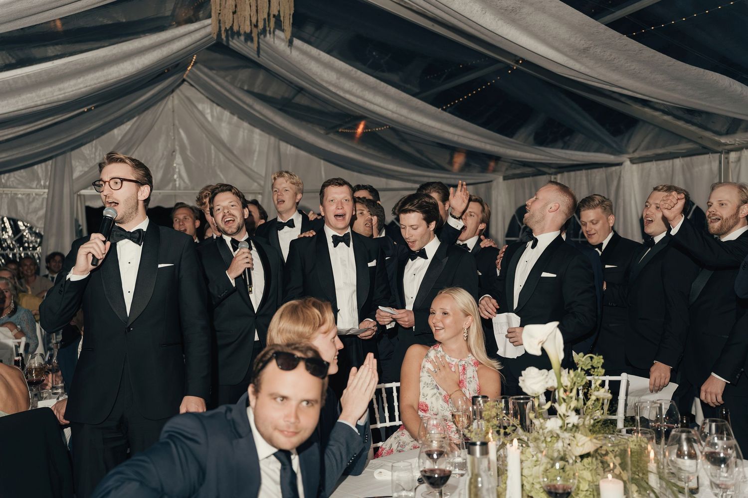 Tuxedo-clad groomsmen cheer and clap around a reception table under a draped tent ceiling at a wedding.