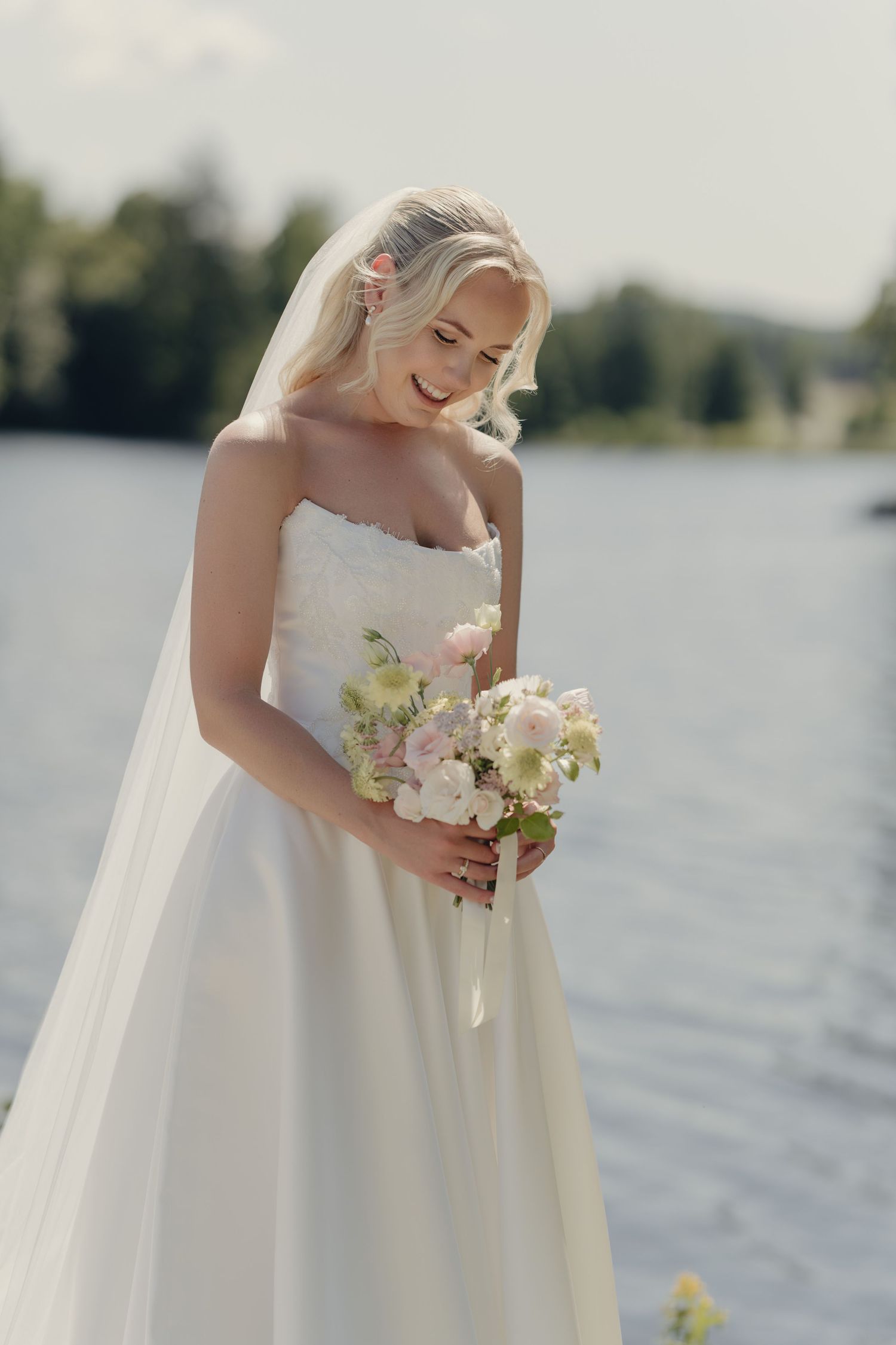 Smiling bride in a strapless white gown and veil holds a pink and white bouquet beside a sunny lake.