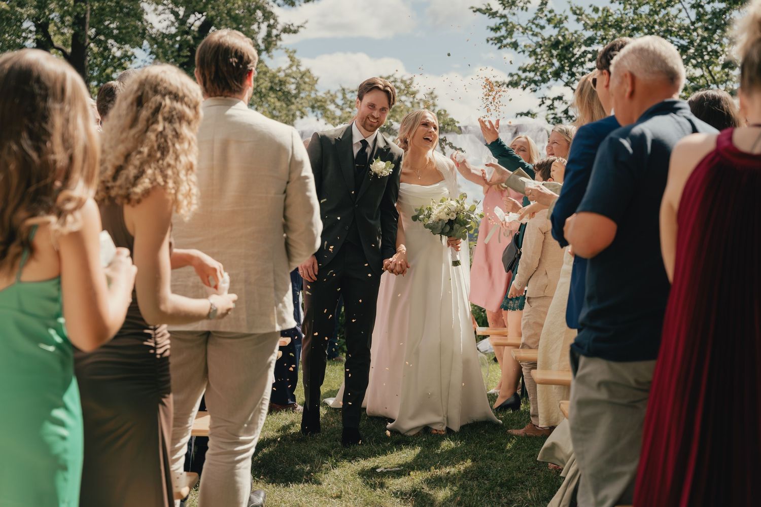 Bride and groom walk through confetti shower surrounded by smiling wedding guests outdoors on sunny day.