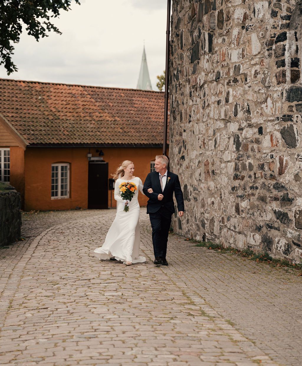 A couple walks along a stone-paved pathway next to a historic stone wall at a wedding venue.