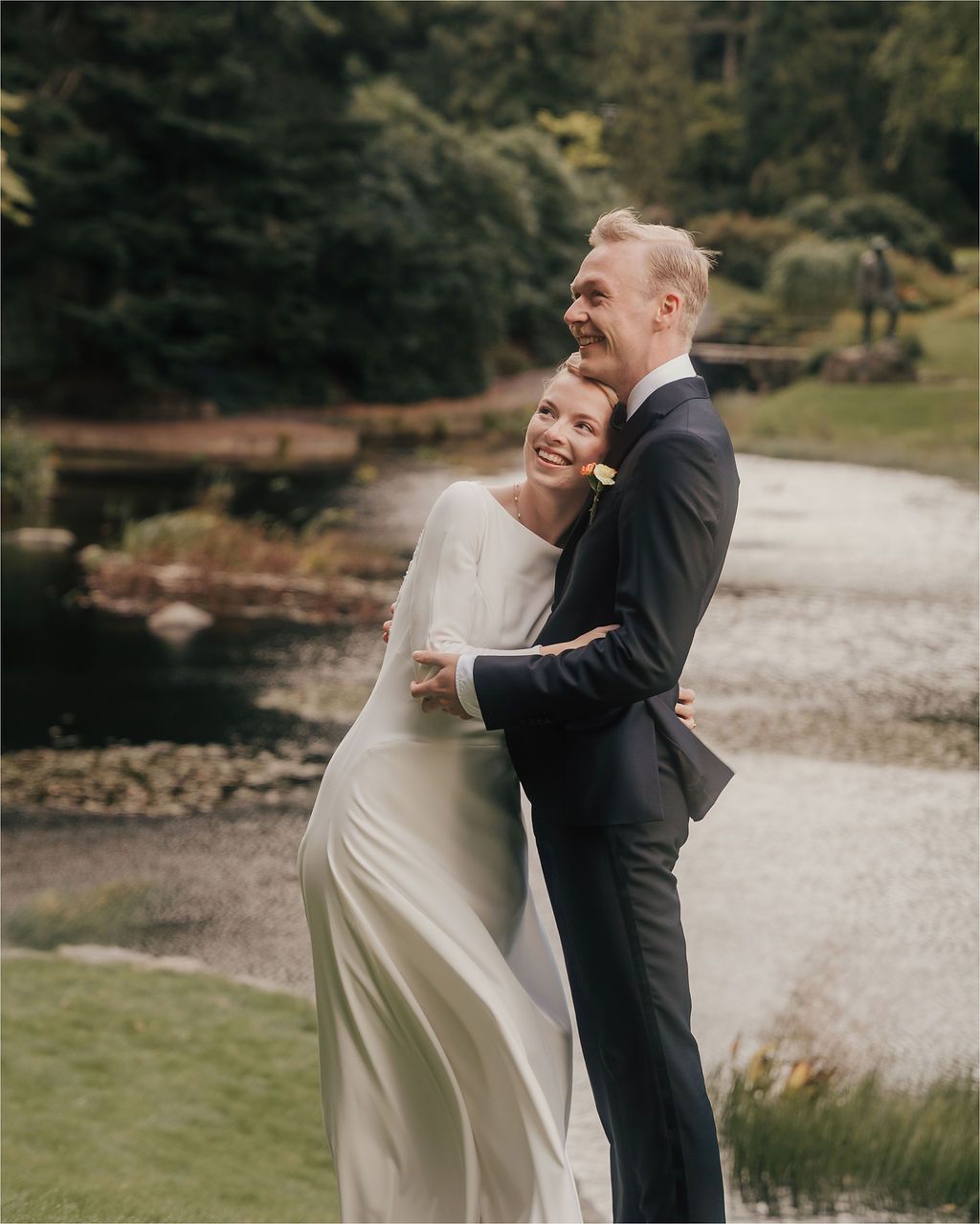 A couple shares a joyful moment together outdoors in formal wedding attire by a scenic waterfront.