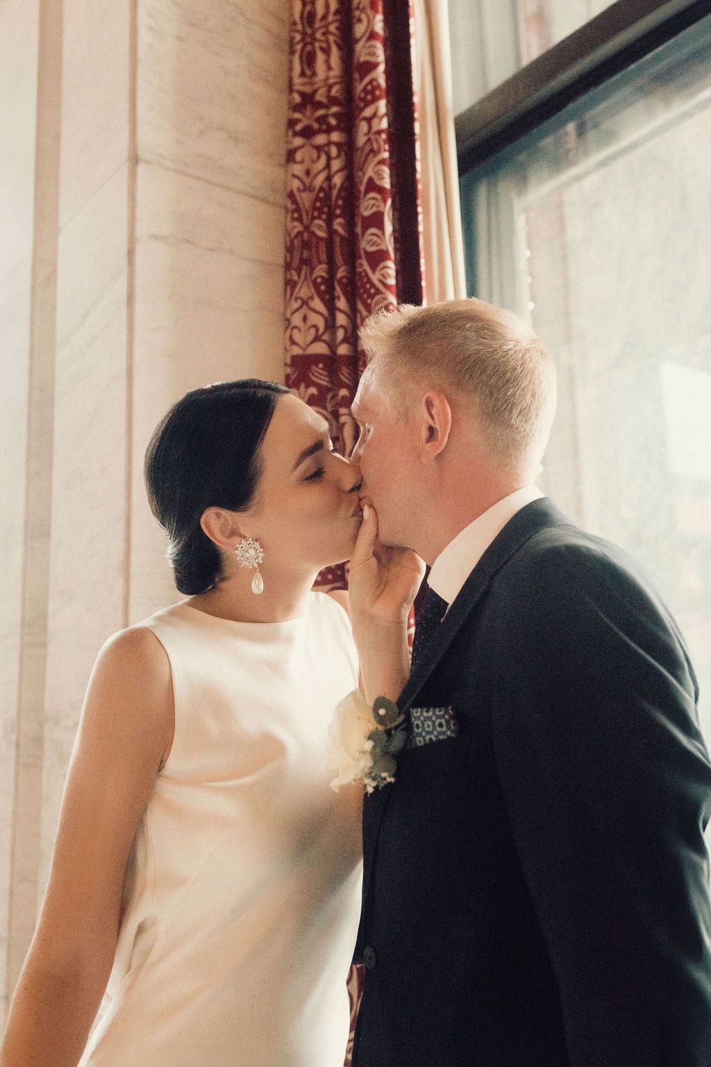 A bride and groom share a romantic kiss in front of elegant curtained windows with soft natural light.