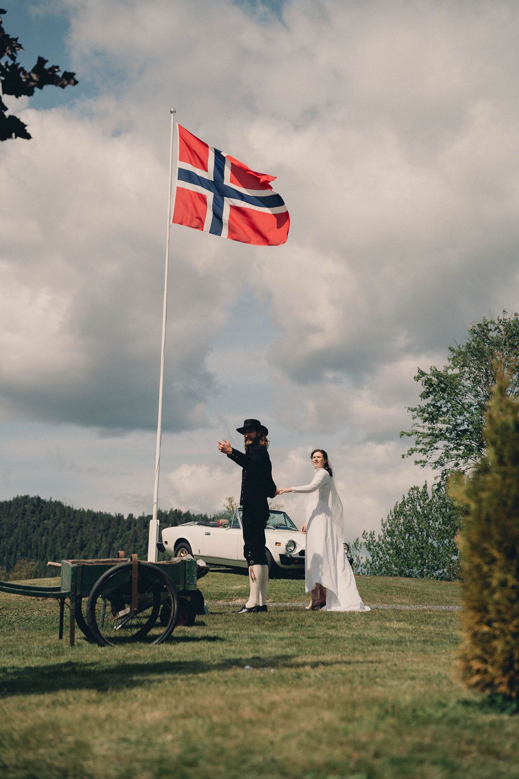 A couple in traditional dress stands beneath a Norwegian flag on a hilltop during what appears to be a wedding ceremony.