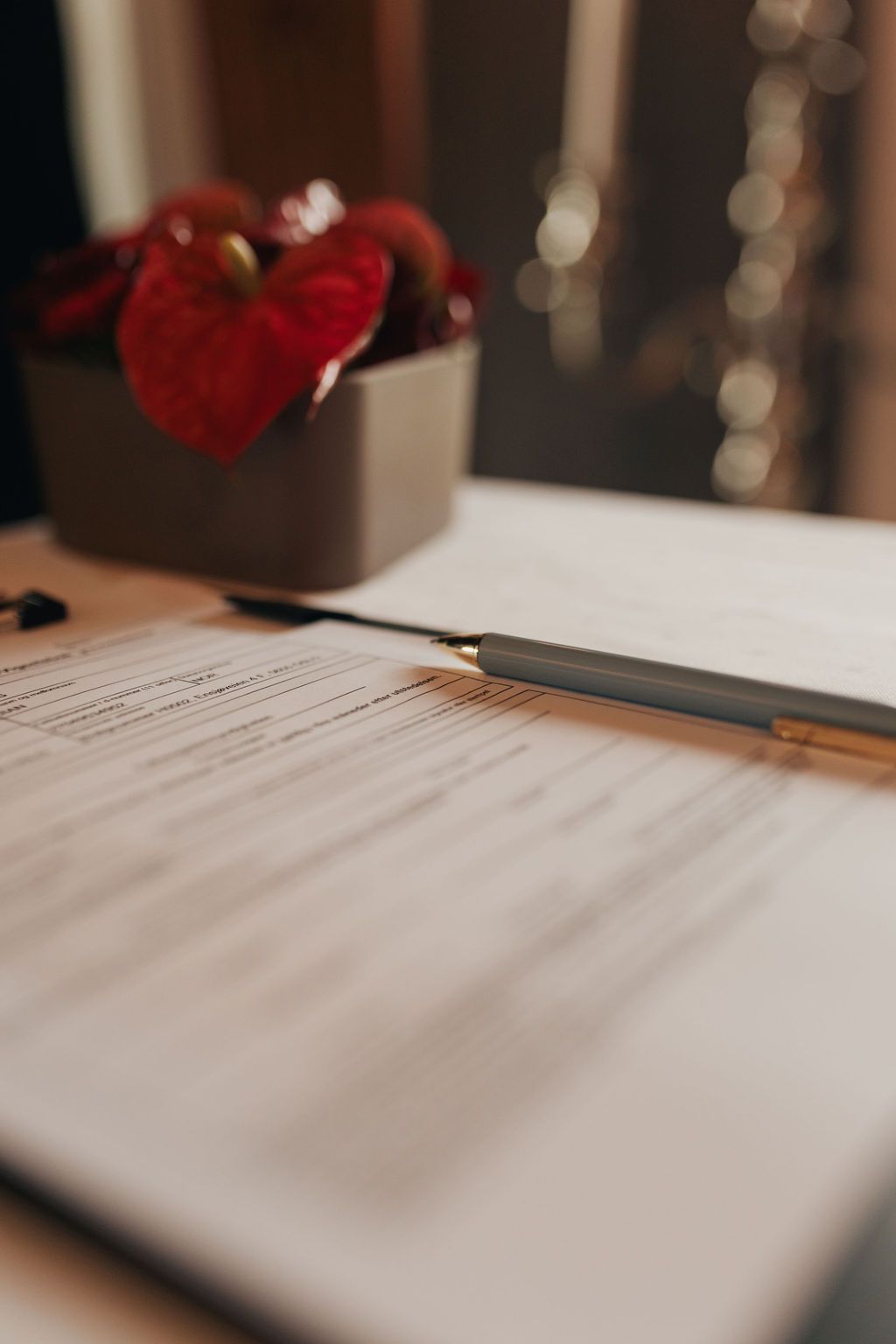 Close-up view of white paper documents on a wooden desk with a red flower accent in soft lighting.