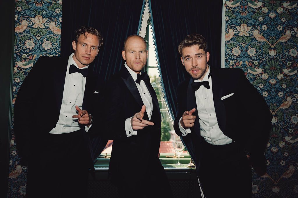 A group in formal black tuxedos and bow ties pose together with drinks at an elegant event.