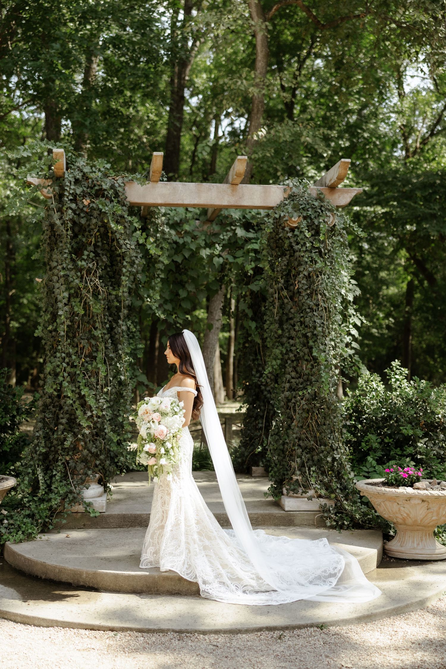 Bride in lace gown and long veil holds bouquet under ivy-covered pergola in lush garden setting.