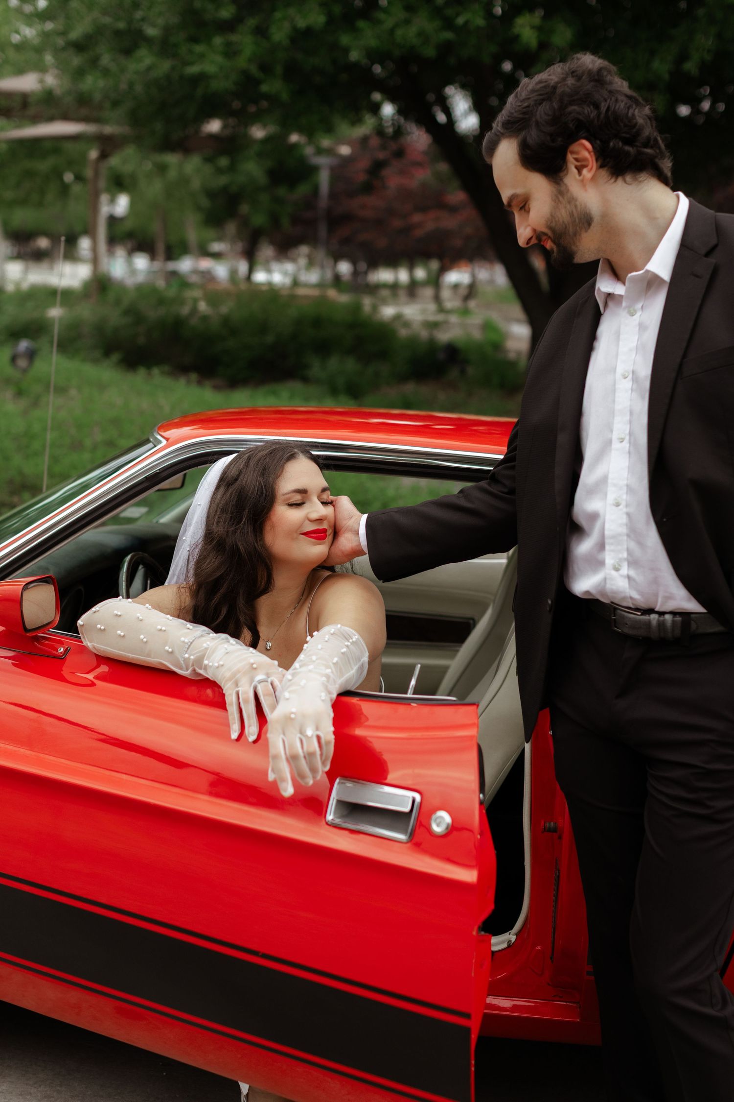 Bride in white gloves leans out of a red classic car while groom in black suit gazes at her.