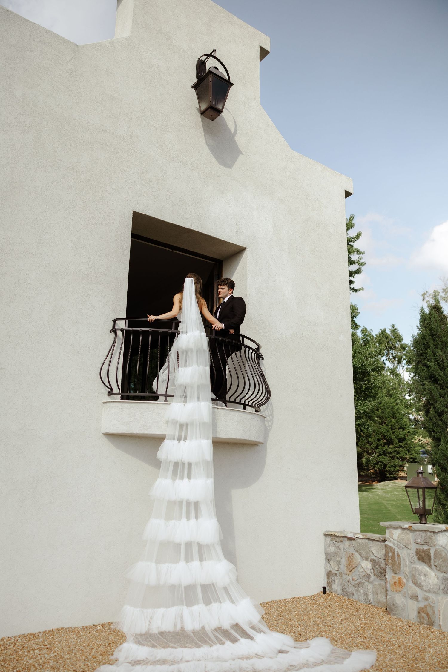 Bride in tiered white veil on balcony with groom in black suit at elegant white stucco venue.