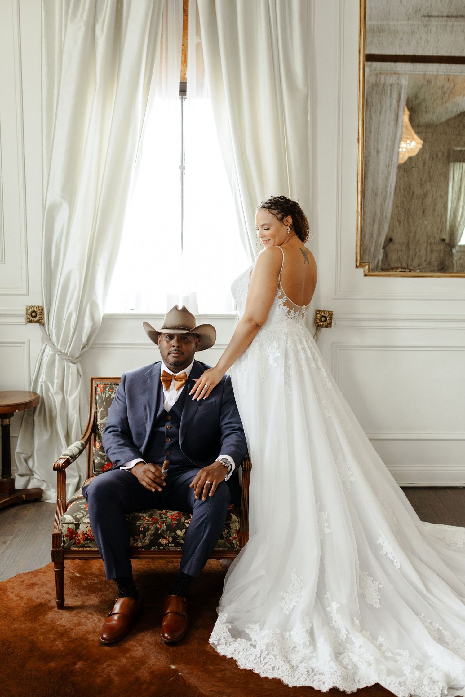 Groom in navy suit and cowboy hat sits in vintage chair while bride in lace backless gown stands beside him.