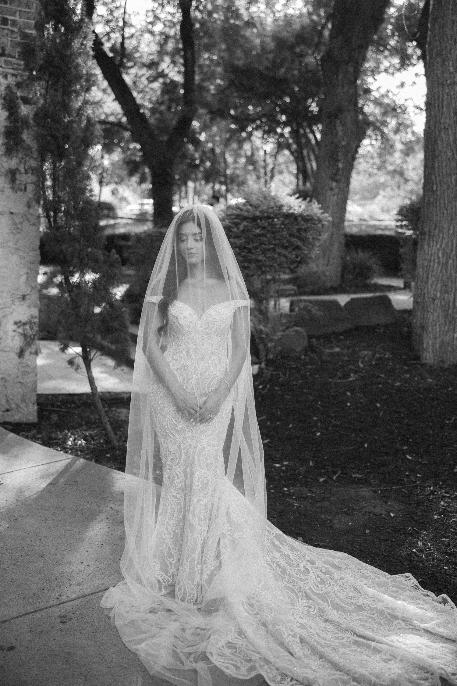 Black and white photo of a bride in a lace gown and flowing veil standing gracefully in an outdoor garden setting.