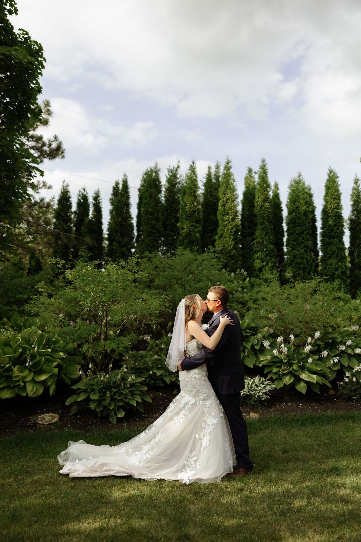 Bride and groom sharing a romantic kiss in a lush garden, with tall evergreen trees and green foliage backdrop.