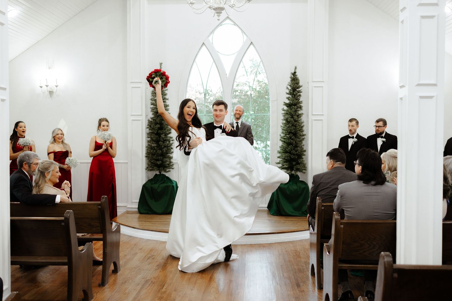 A joyful wedding couple celebrates inside a charming white chapel with gothic windows and wooden floors.