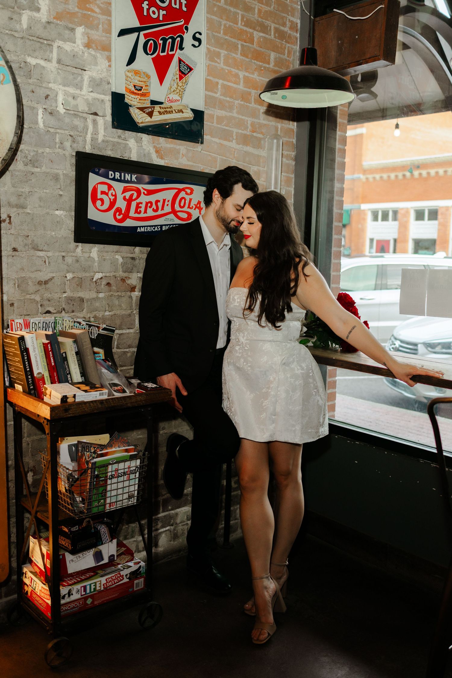 A couple in wedding attire embraces near a window in a vintage diner with retro Pepsi-Cola and Tom's signs on brick walls.