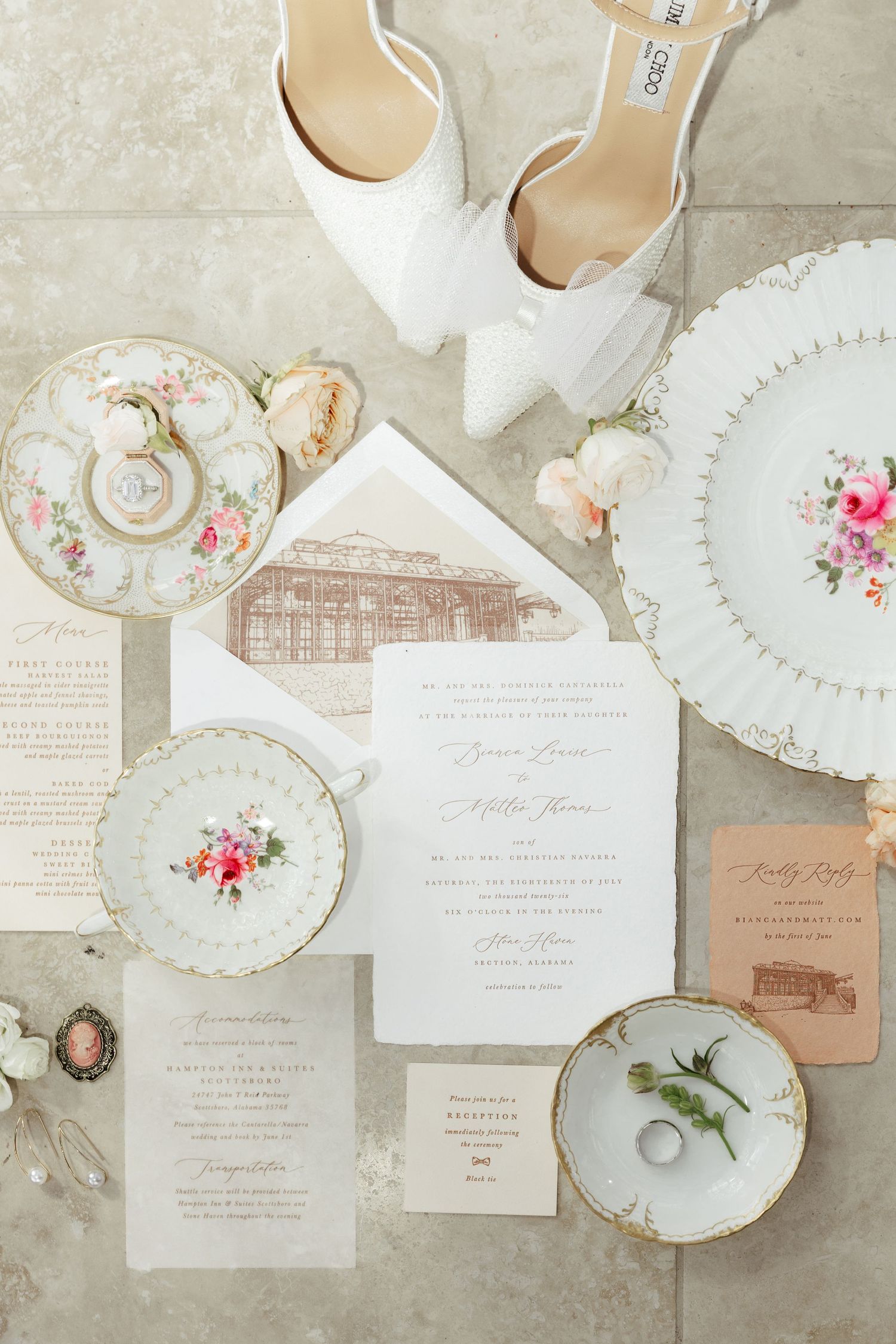 Elegant wedding flat lay with floral china, white lace heels, invitation suite, and roses on a neutral stone surface.