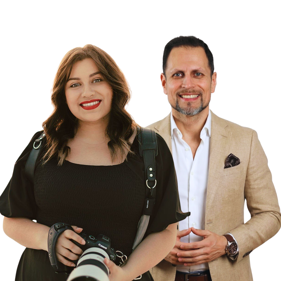 Professional photographer in black dress holding camera poses with colleague wearing beige blazer against white background.