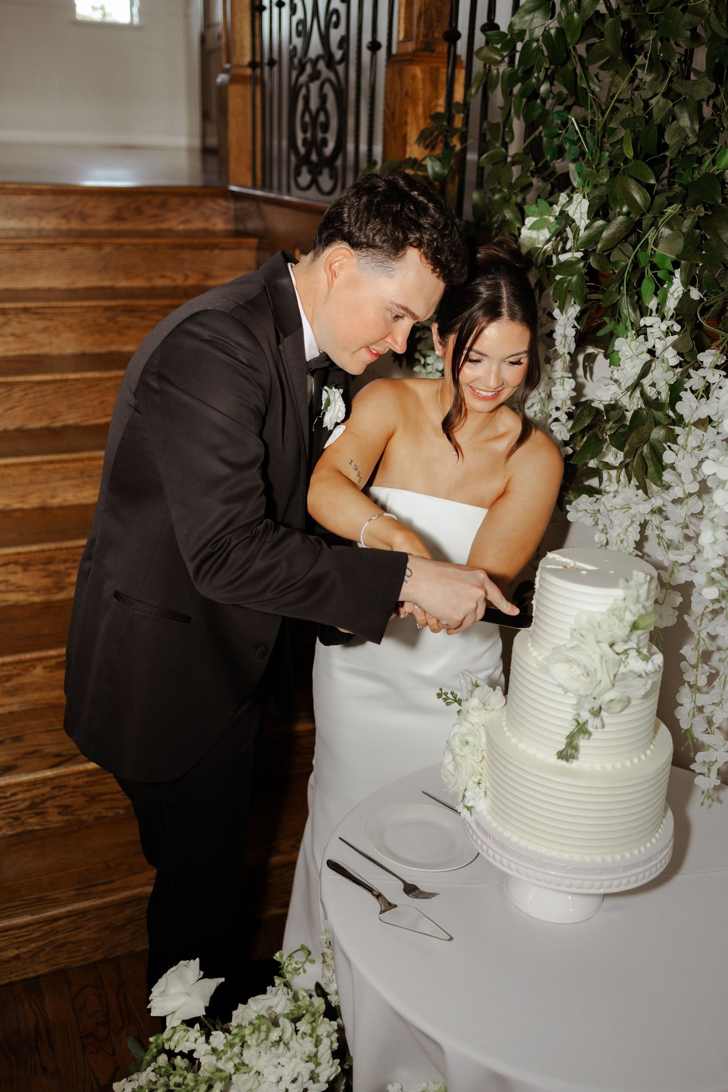 Bride and groom cutting white tiered wedding cake near floral greenery backdrop and wooden staircase.