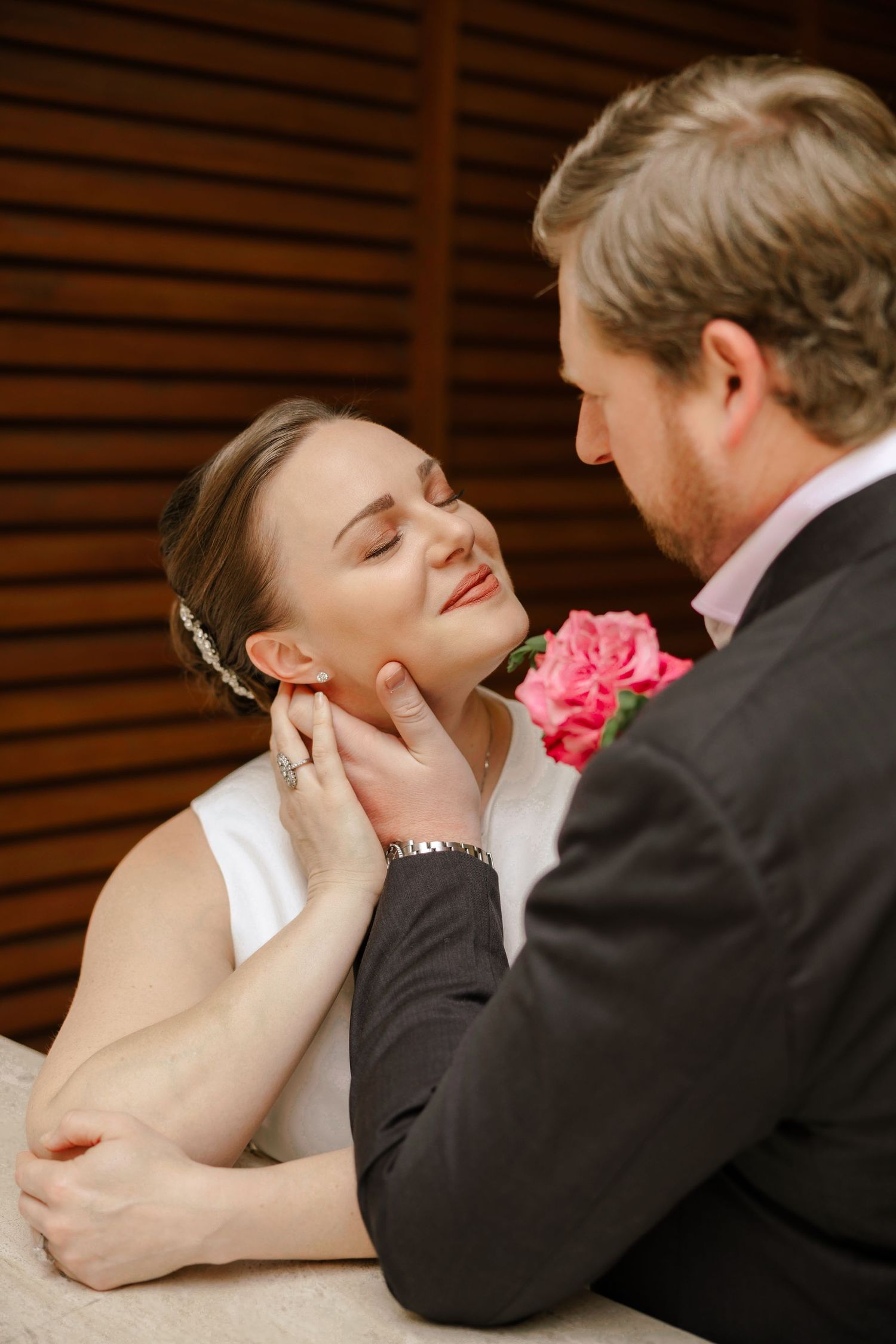 A groom tenderly cups his bride's face as she smiles with closed eyes, wearing a white dress and floral hair accessory.