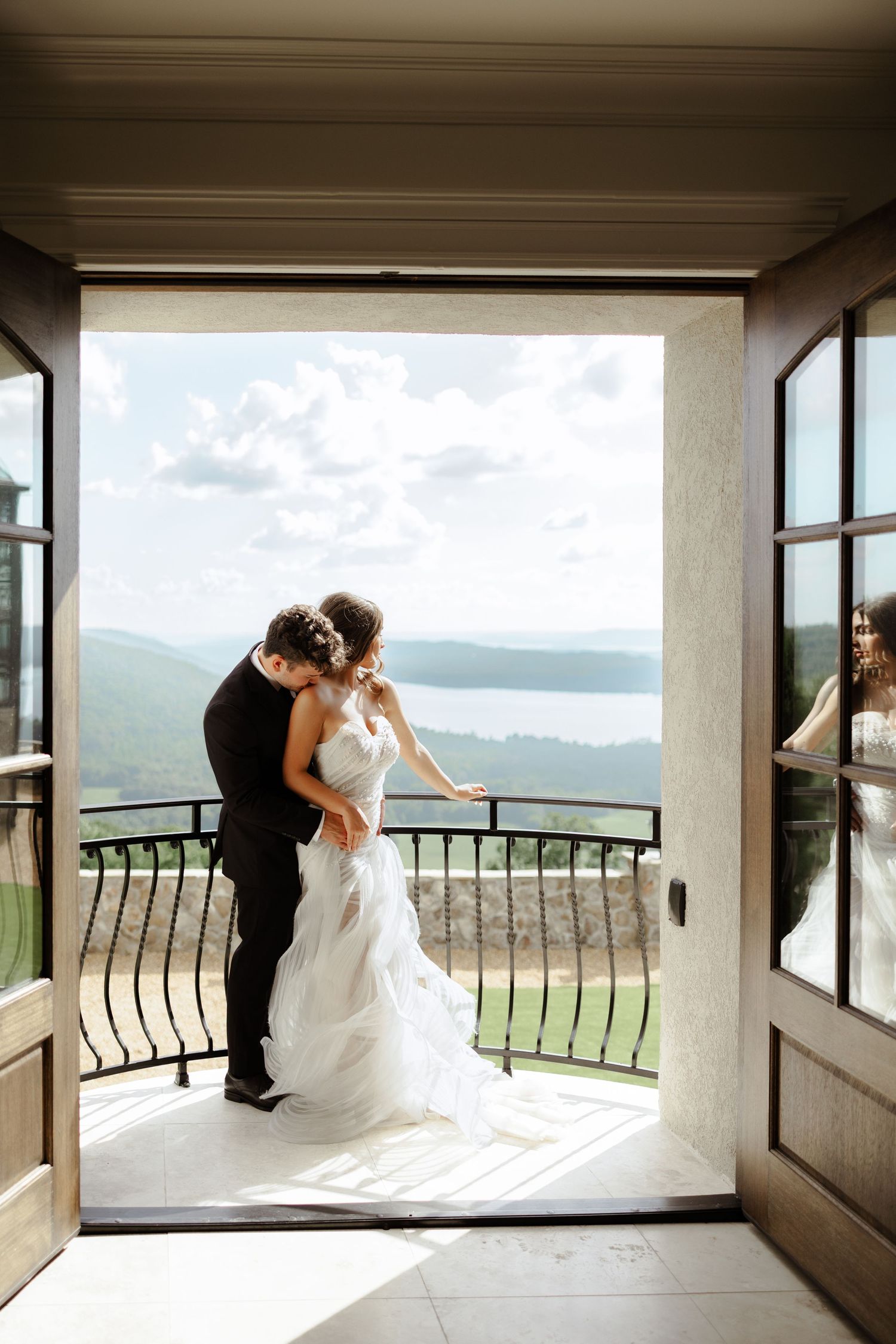A bride and groom share a romantic embrace on a balcony with scenic mountain and lake views through open French doors.
