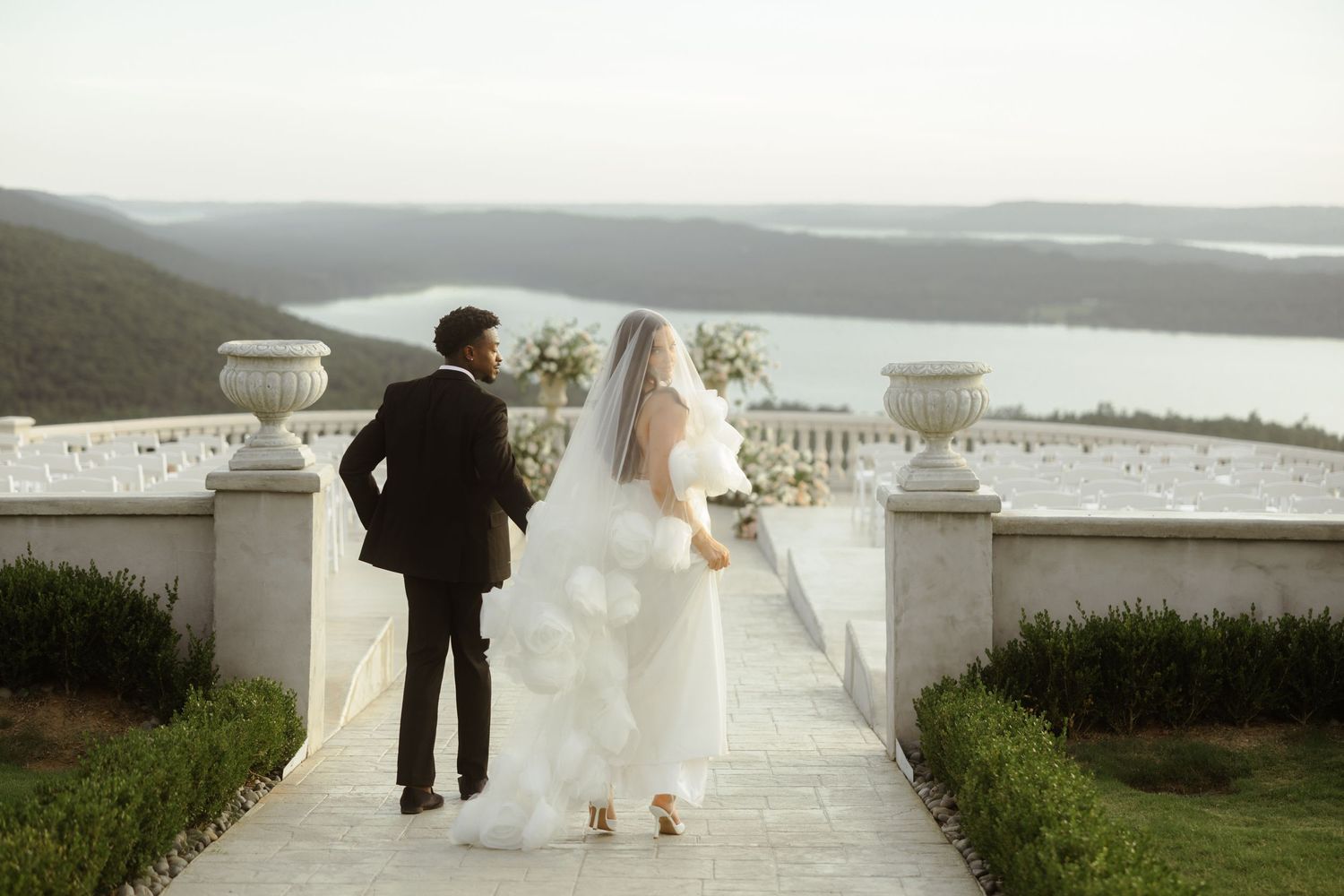 Bride and groom walk hand in hand on elegant terrace overlooking scenic lake and rolling hills at outdoor wedding.
