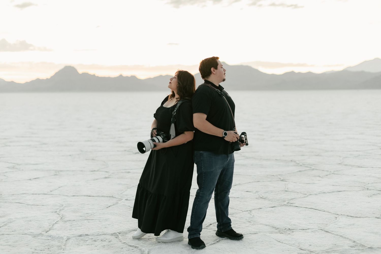 Husband & wife wedding photo/video team at Bonneville salt flats, at sunset with mountains in the distance and cameras in hand.