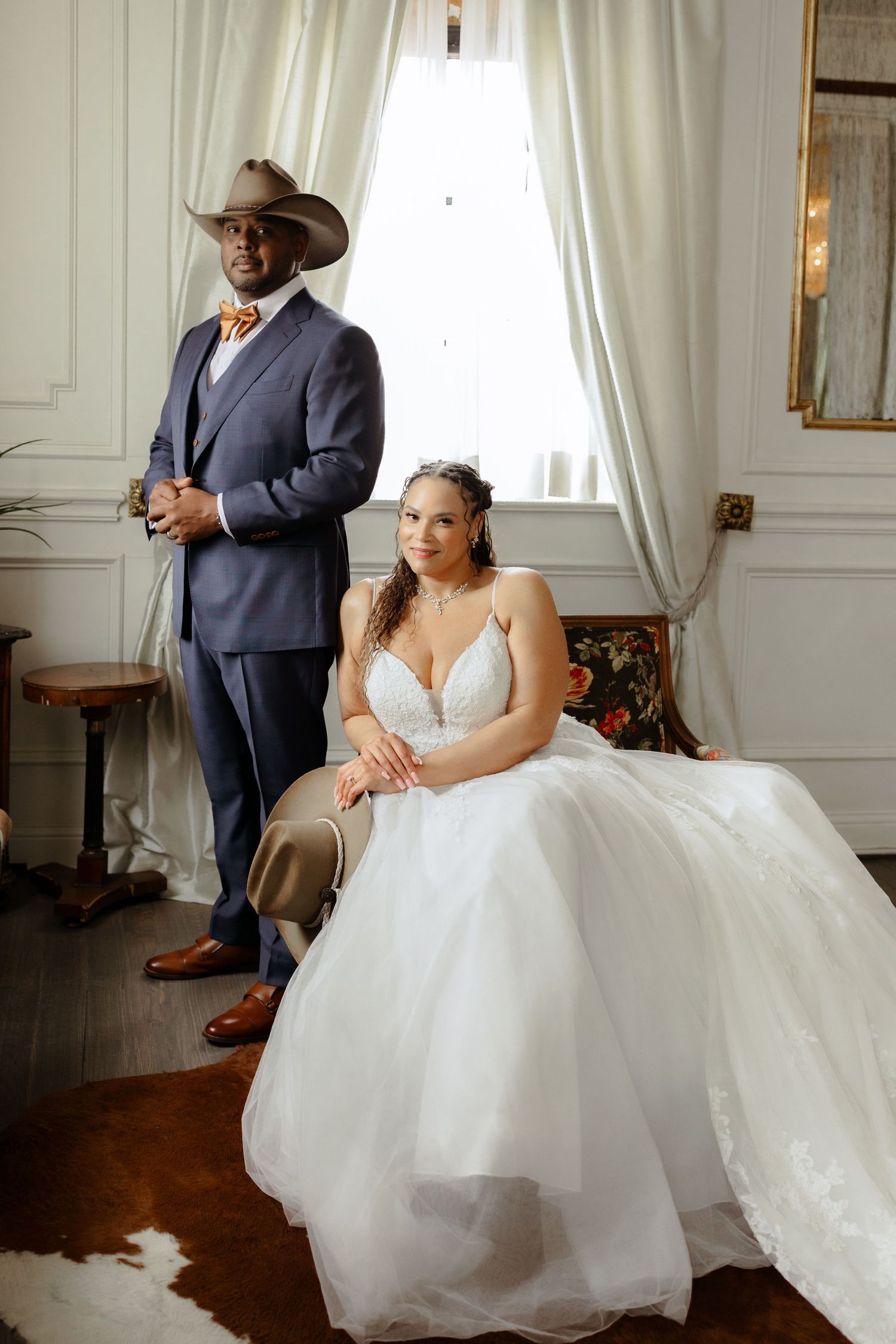 Bride in white ballgown sits beside groom in navy suit and cowboy hat near window in elegant white room.