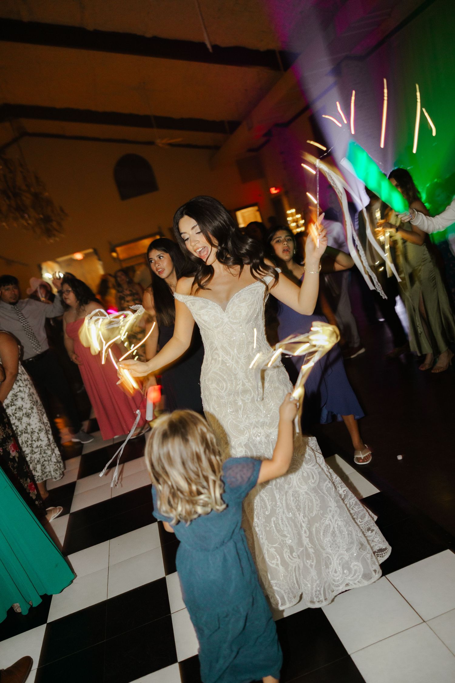 Bride in white lace gown dances with child on checkered floor at wedding reception with glowing wands.