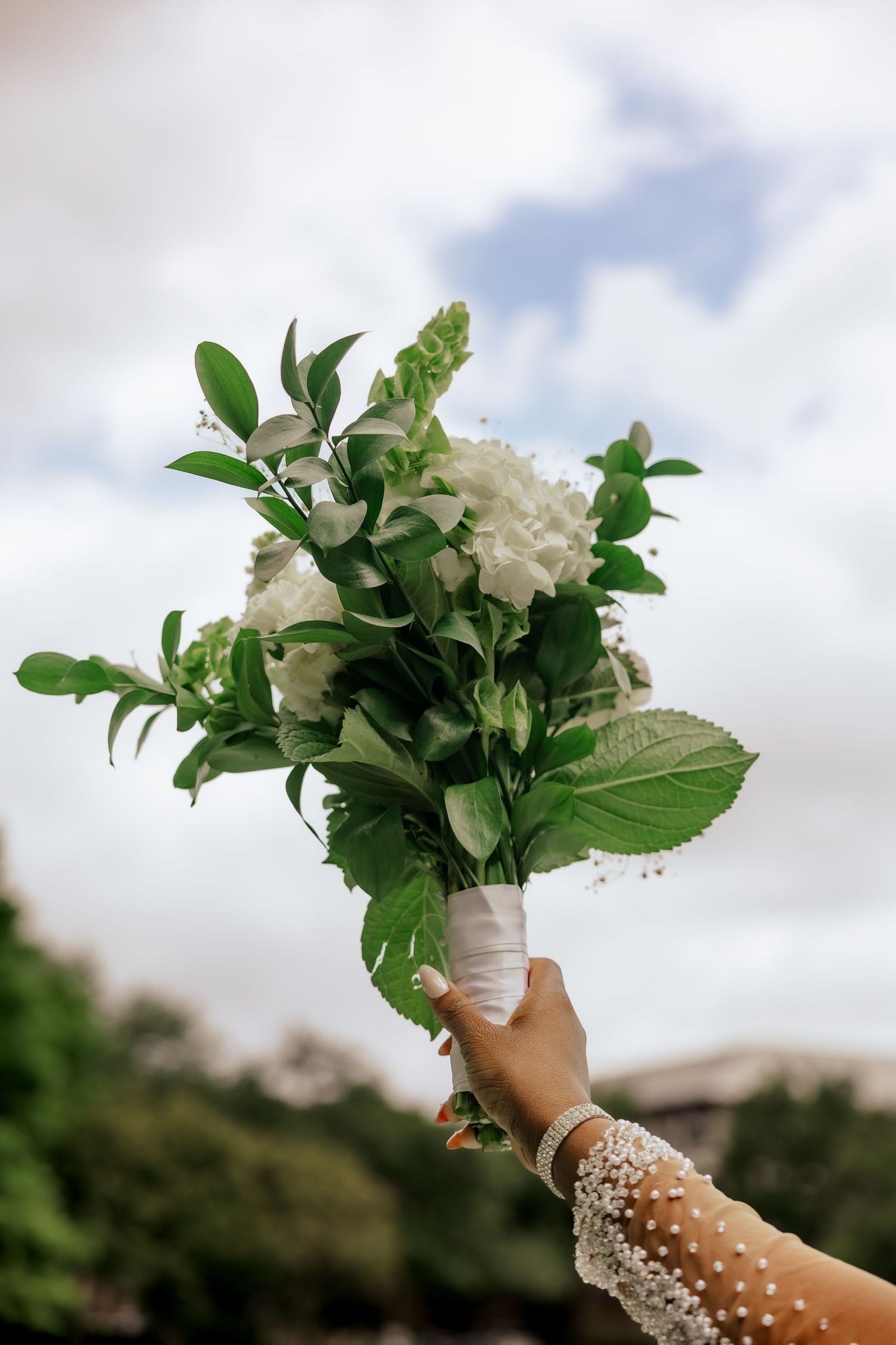 A hand adorned with pearl bracelets holds a white floral bouquet with green leaves against a cloudy sky backdrop.