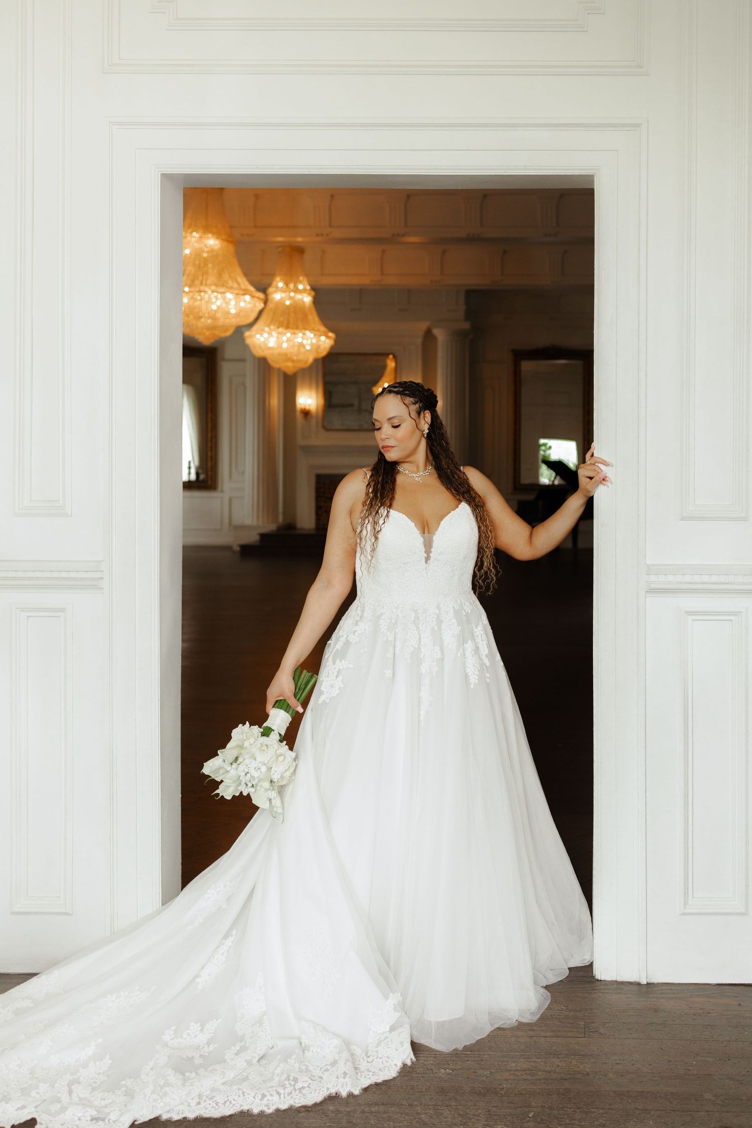 Bride in a white lace A-line wedding gown holding a bouquet, standing in an elegant doorway with chandeliers.