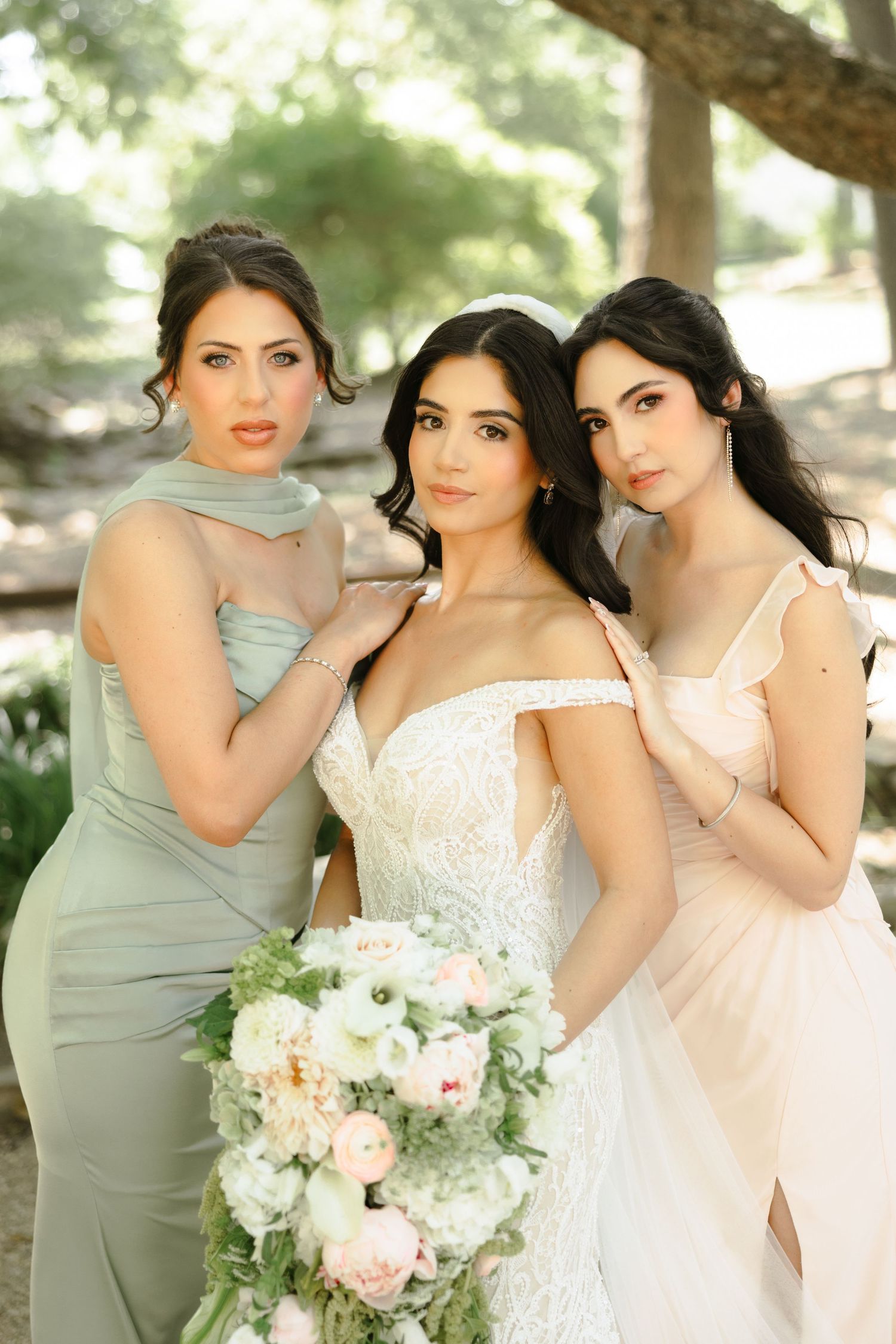Bride in white lace gown holds floral bouquet flanked by two bridesmaids in sage and blush dresses outdoors.