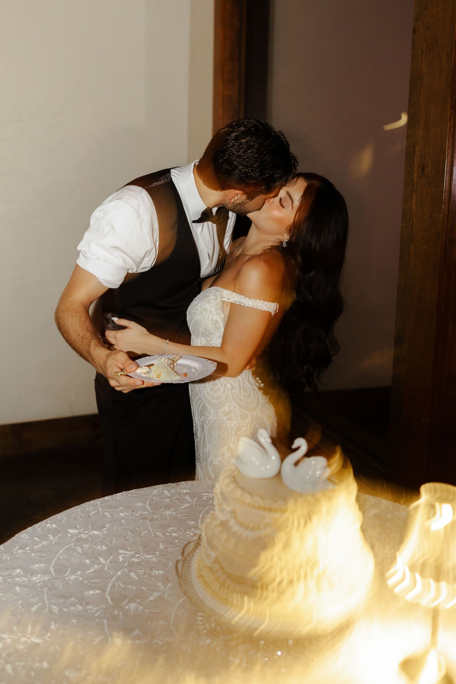 Bride and groom sharing a kiss while cutting their white wedding cake adorned with swan toppers at their reception.