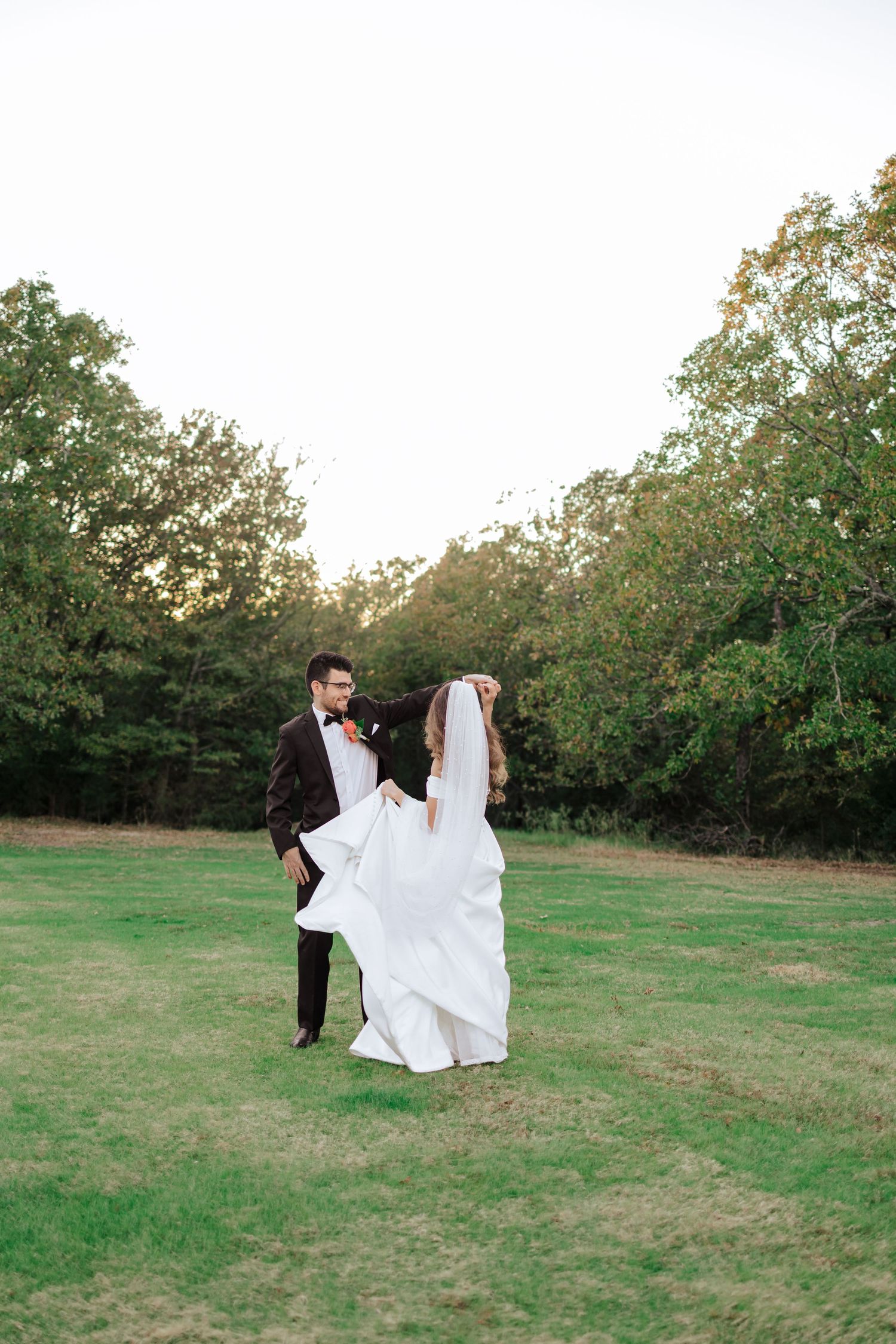 A couple in wedding attire dances together on a green lawn surrounded by trees at sunset.