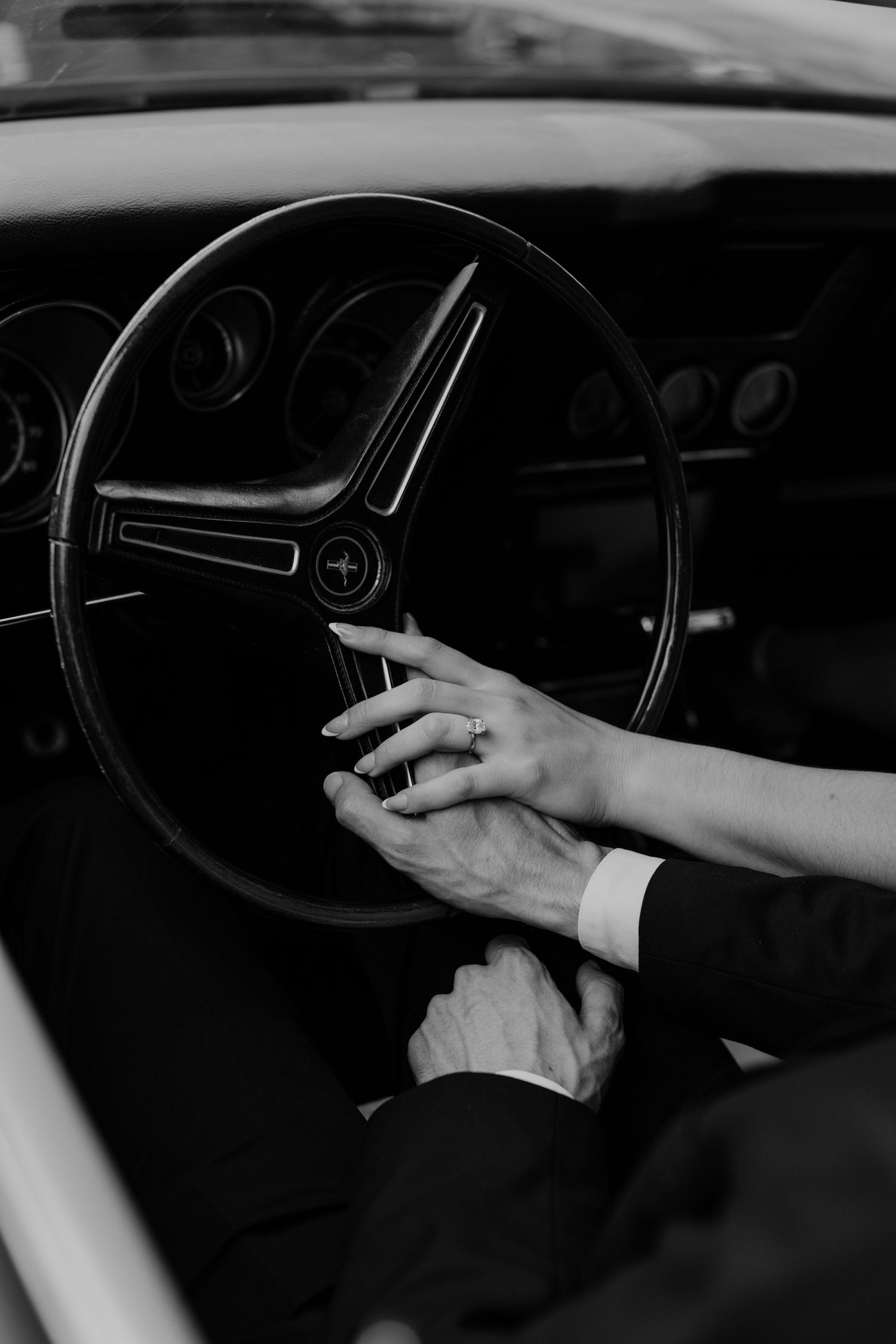 Black and white photo of a couple holding hands on a vintage Ford Mustang steering wheel, wearing wedding rings.