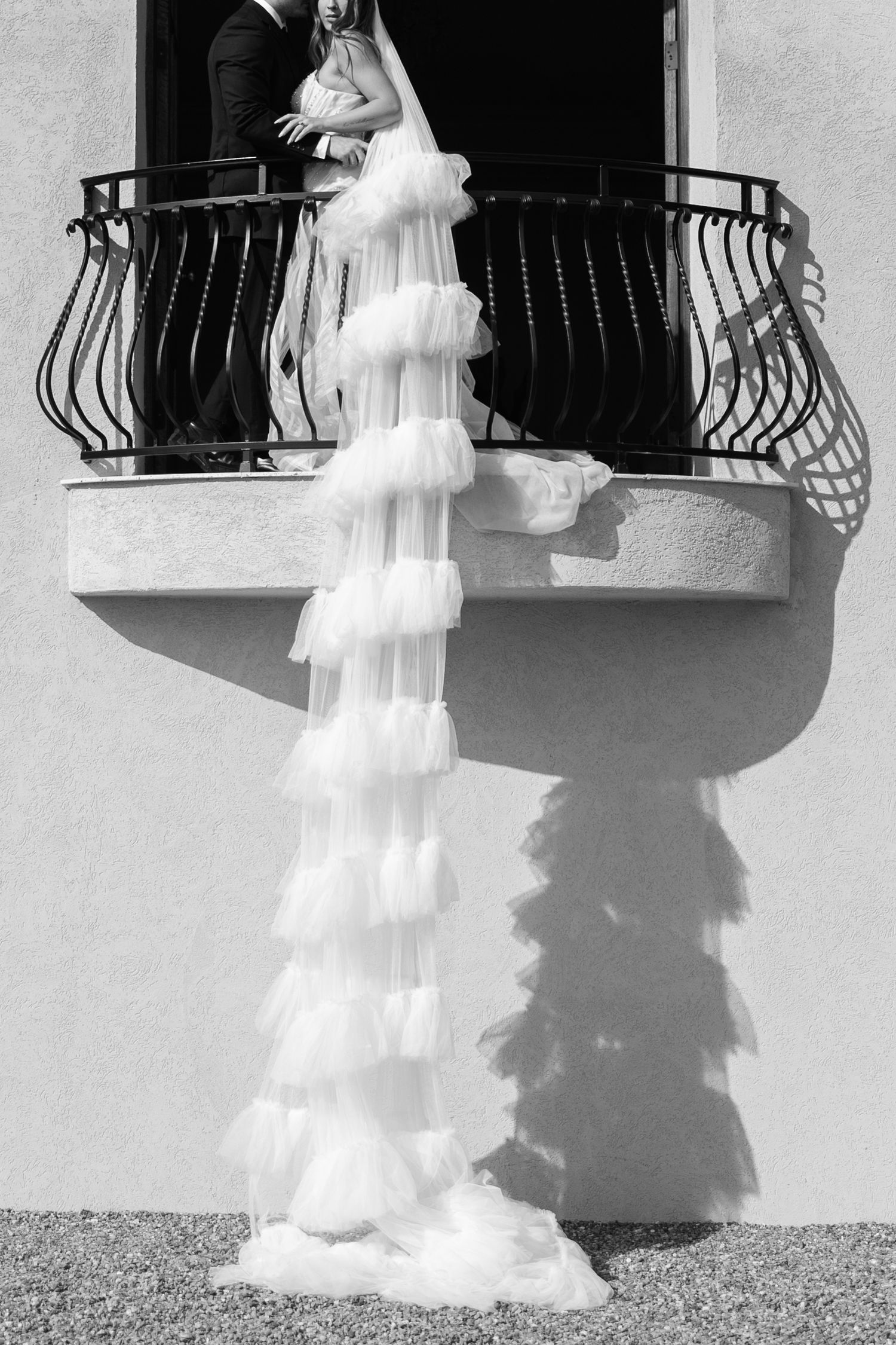 Black and white wedding photo of bride in ruffled gown cascading from balcony where couple embraces.