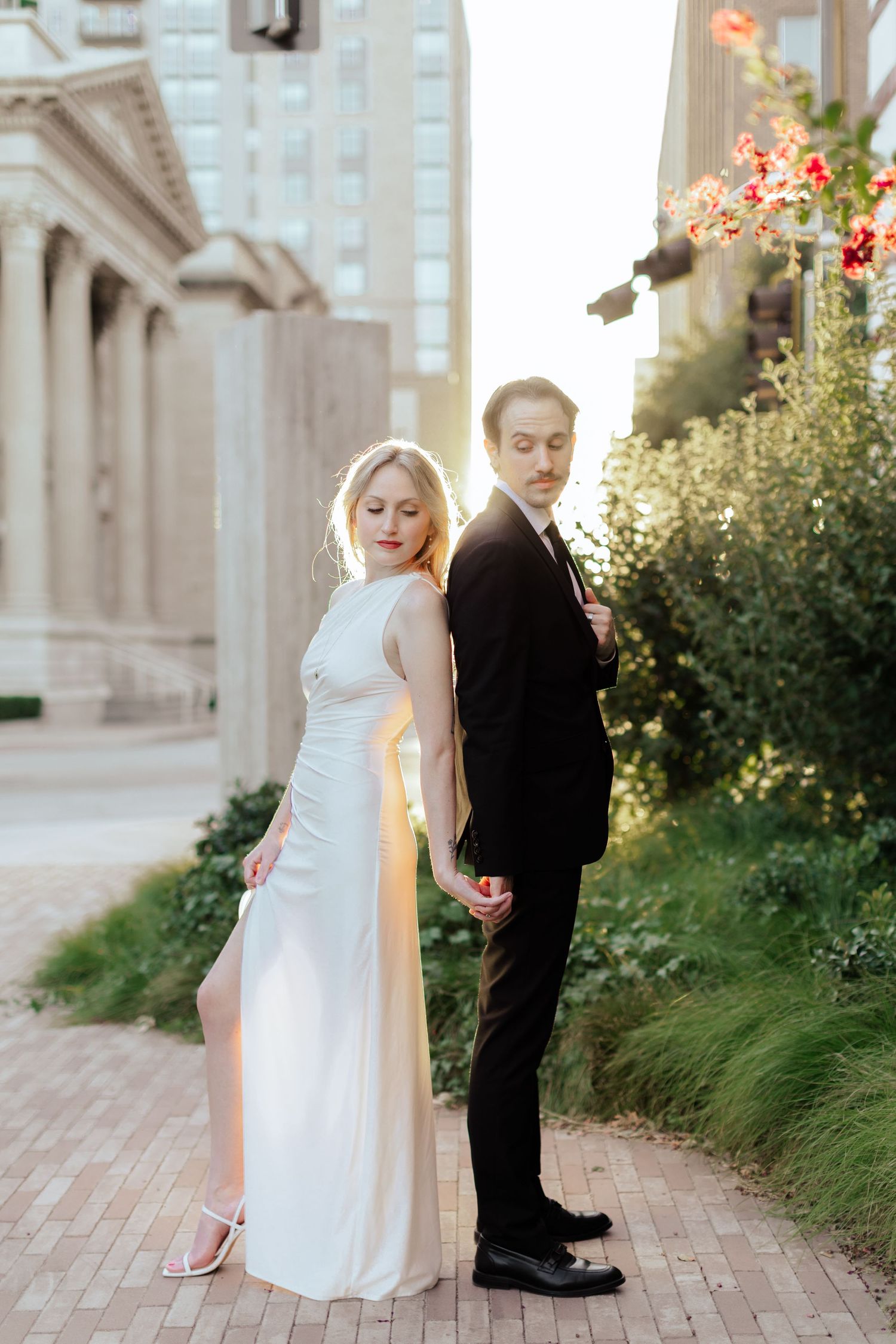 Wedding couple poses playfully on brick pathway surrounded by elegant architecture and flowering bushes.