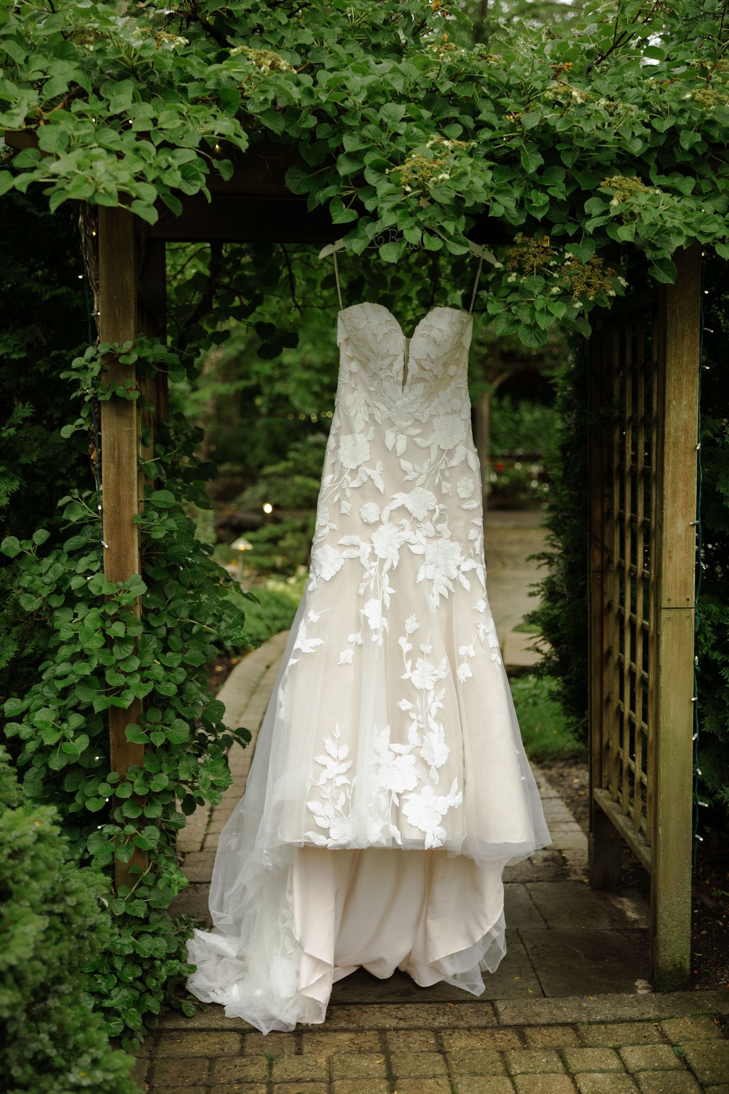 Elegant strapless mermaid wedding dress with floral appliques hanging in a lush green garden arbor.