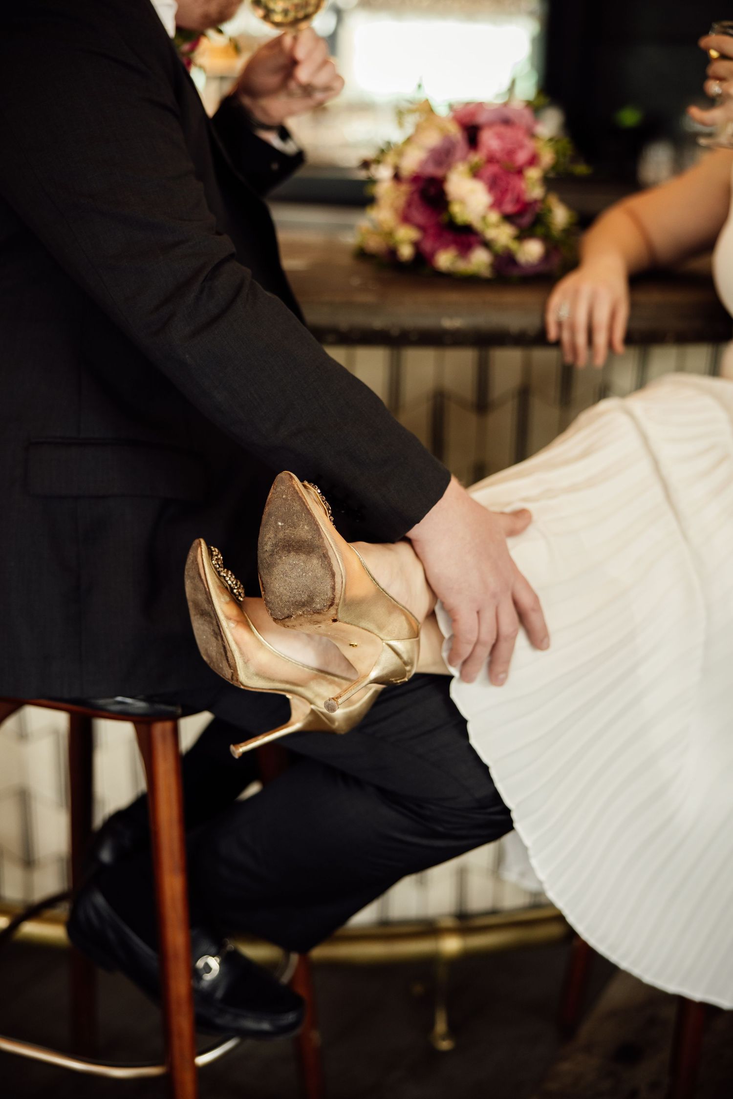 Bride in white dress with gold heels resting on groom's lap in black suit at wedding reception.