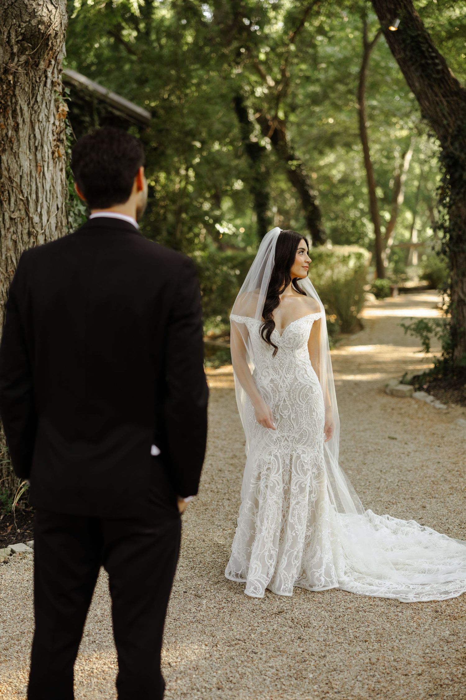Groom watches bride in lace mermaid wedding dress and veil smile on a tree-lined garden path.