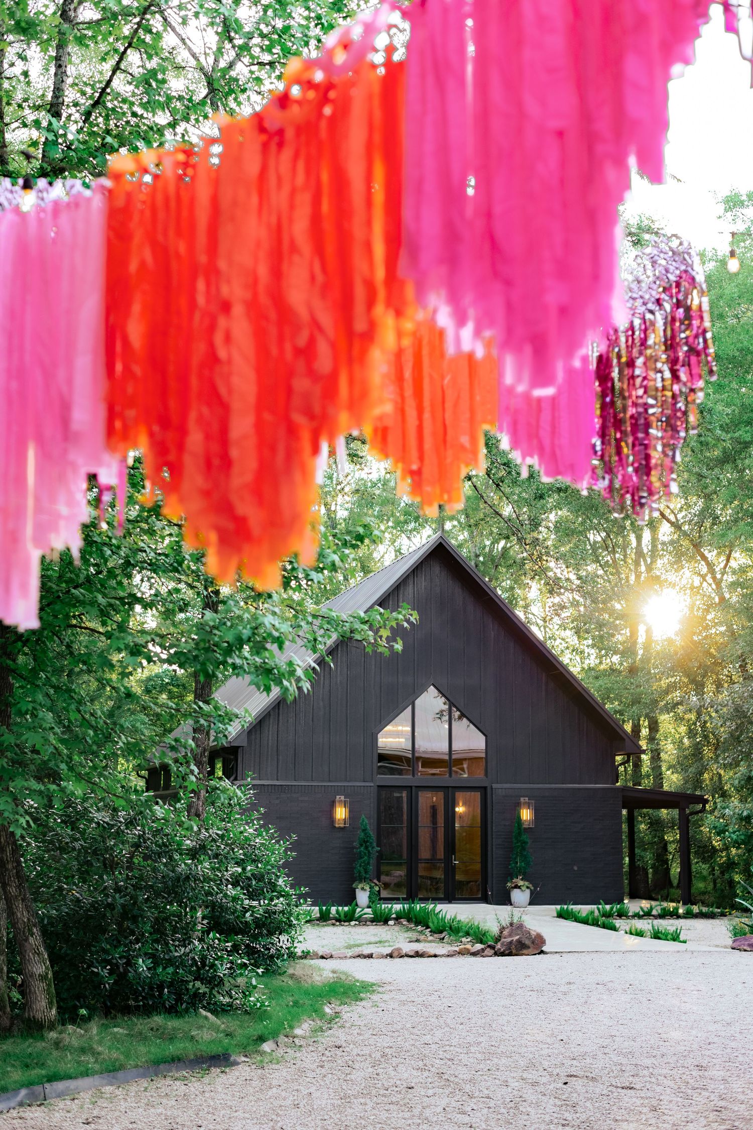 Modern black barn venue with vibrant pink and orange tissue paper decorations hanging against a sunset-lit forest backdrop.
