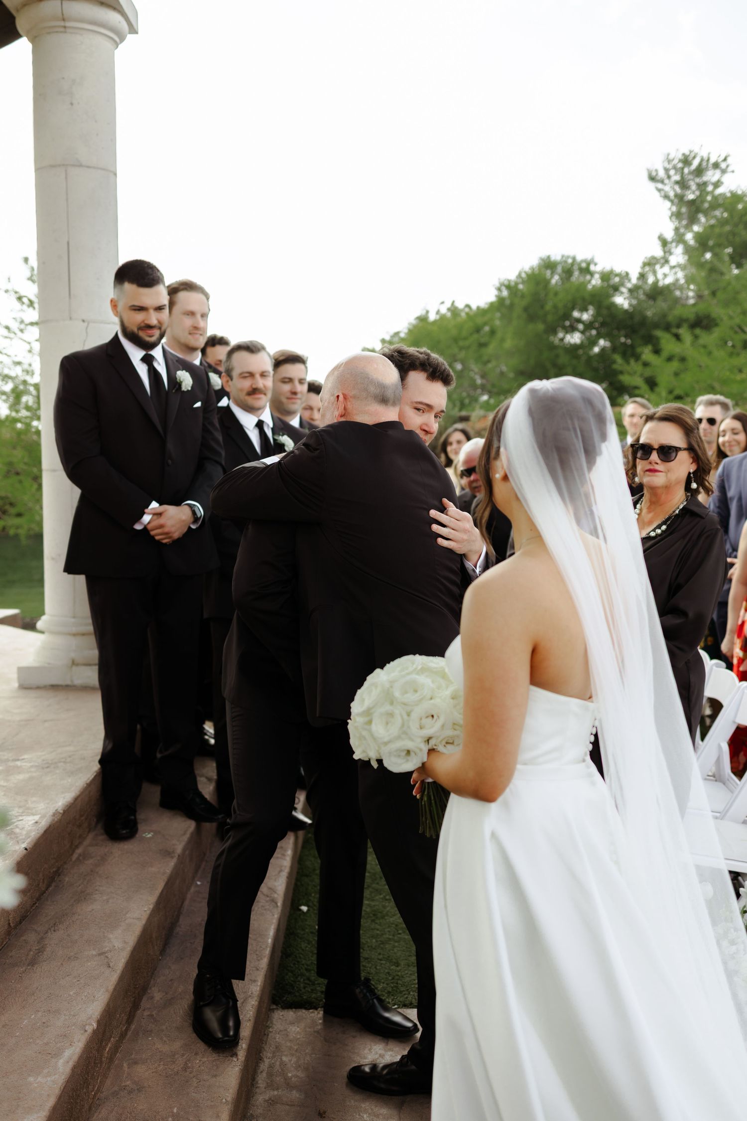 Bride in white gown holding bouquet approaches groom and groomsmen in black suits at outdoor wedding ceremony.