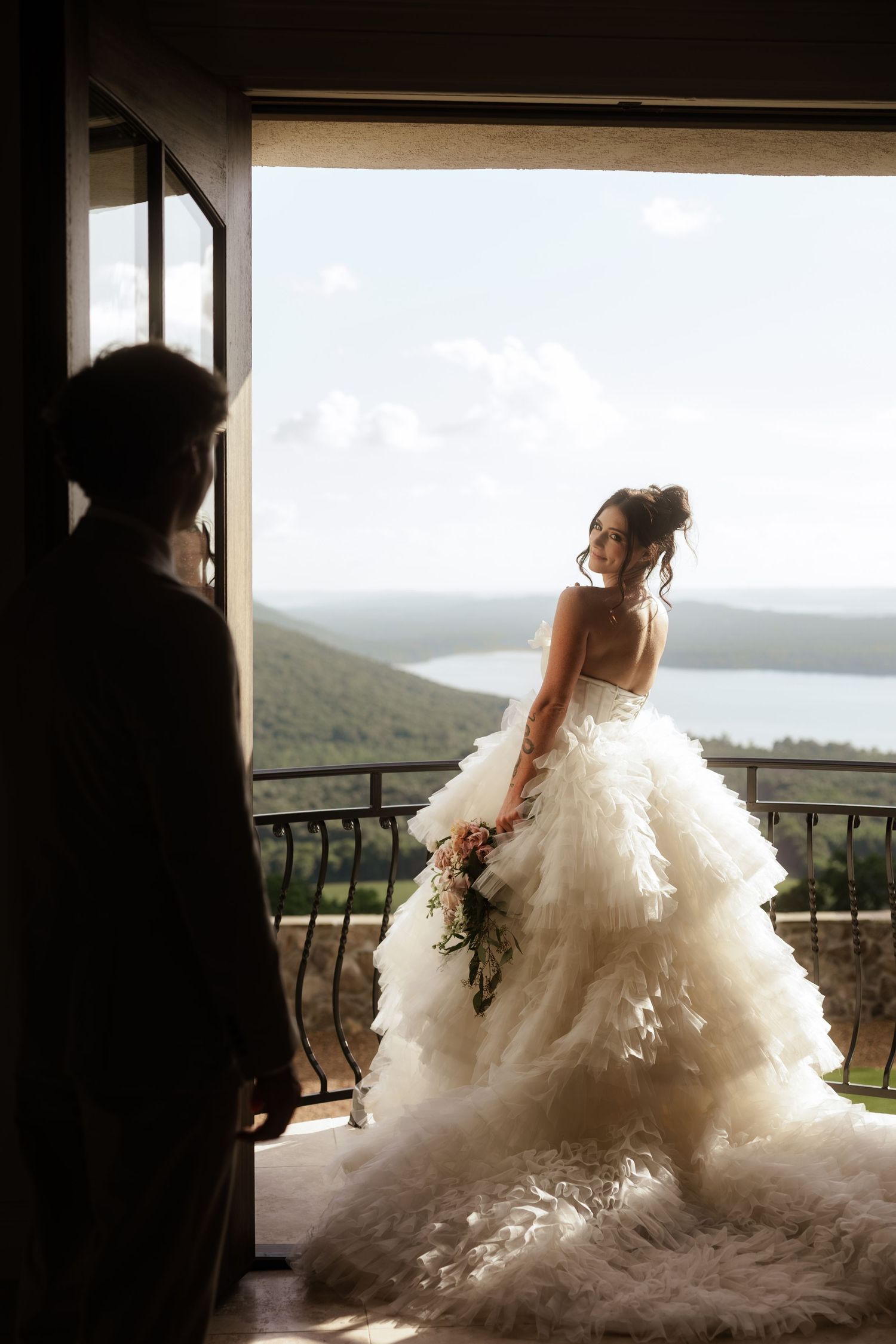 Bride in a ruffled white gown holds bouquet on a balcony overlooking a lake, as groom watches from doorway.