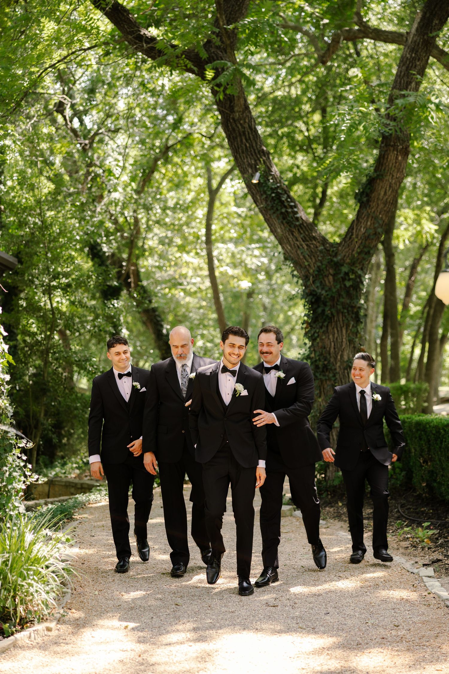Five groomsmen in black tuxedos with bow ties walk confidently down a sunlit garden path surrounded by lush green trees.