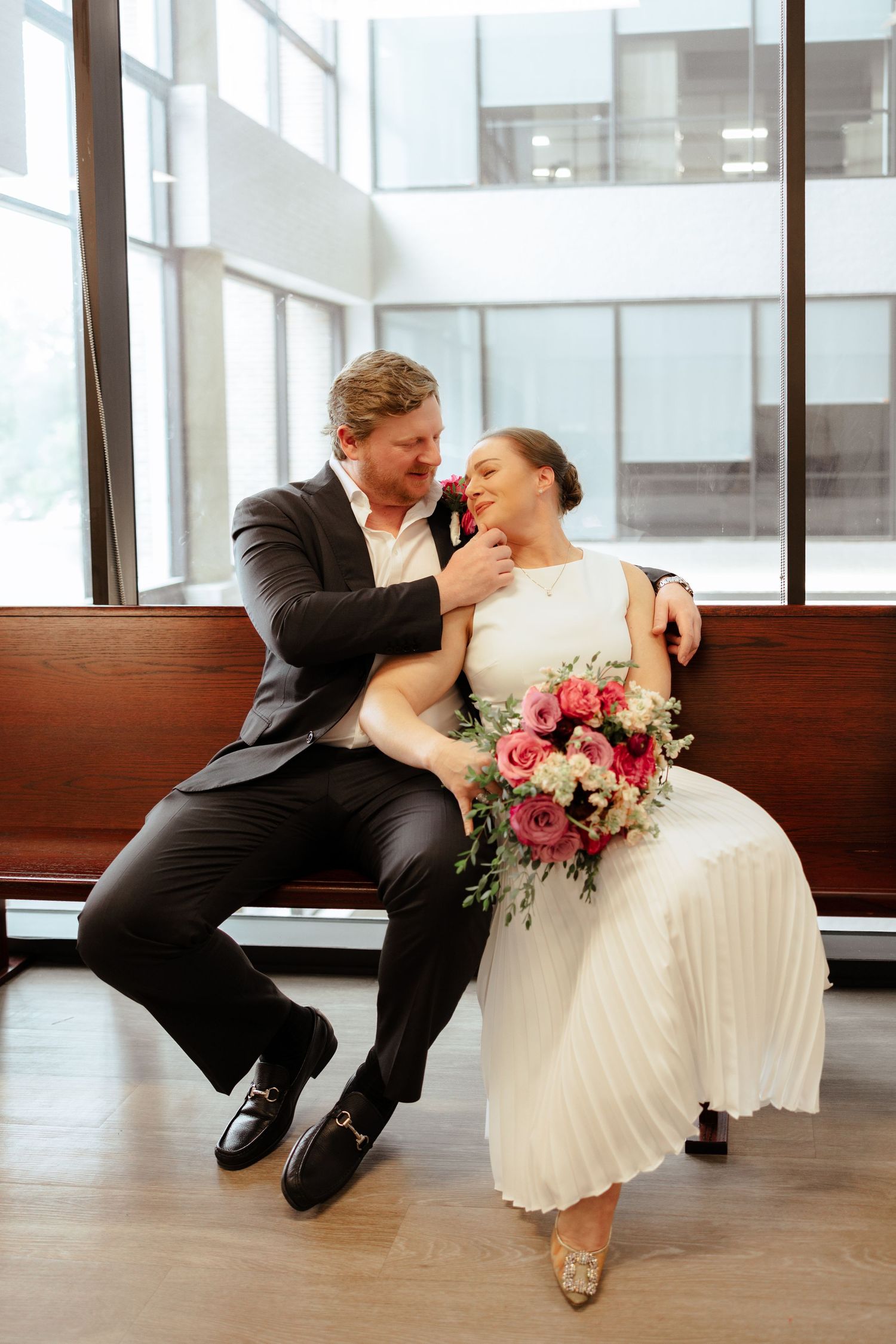 Bride and groom sit closely on a bench, sharing a tender moment, bride holding a pink and white bouquet.