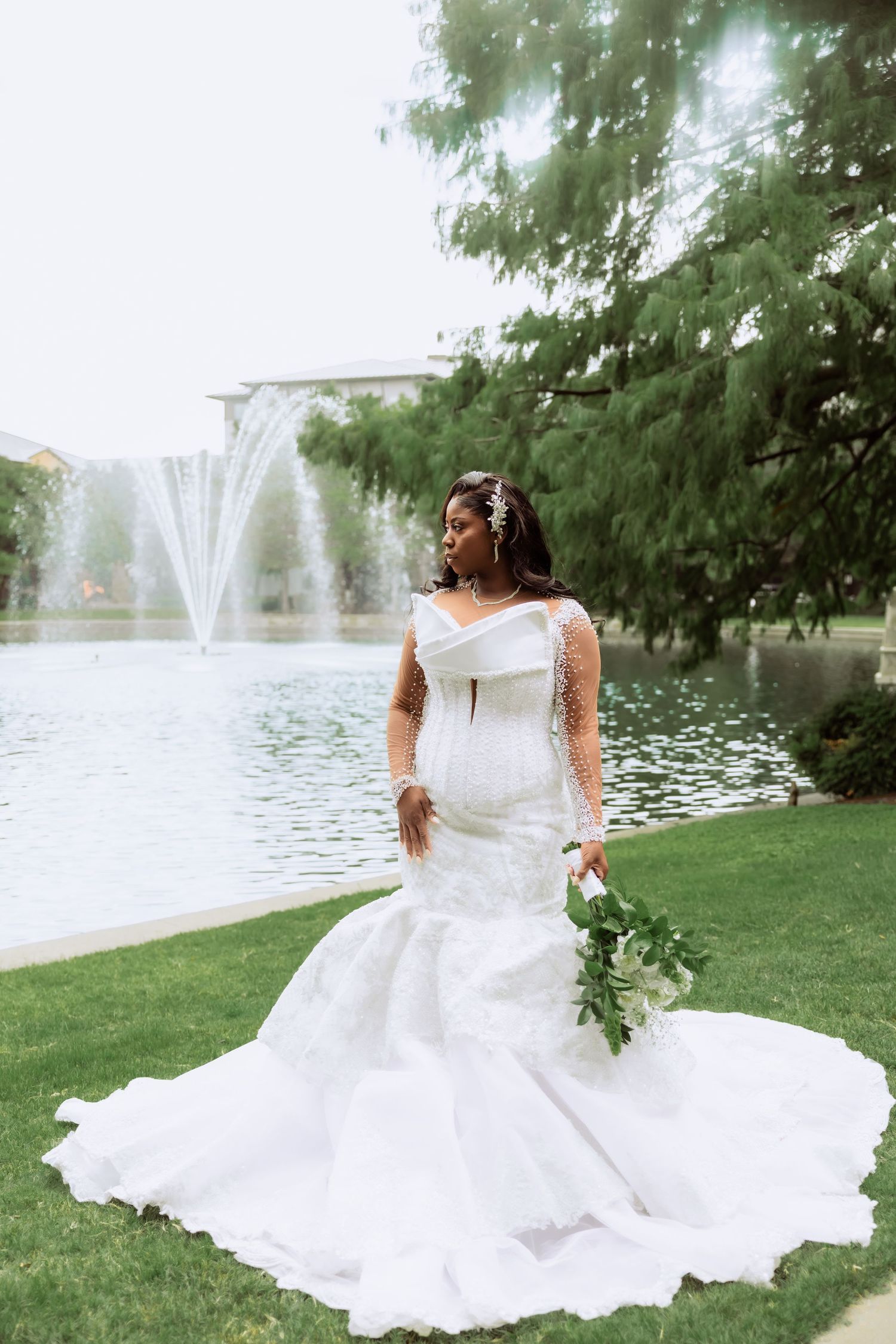 A bride in a flowing white wedding dress poses gracefully by a lake with fountains in the background.