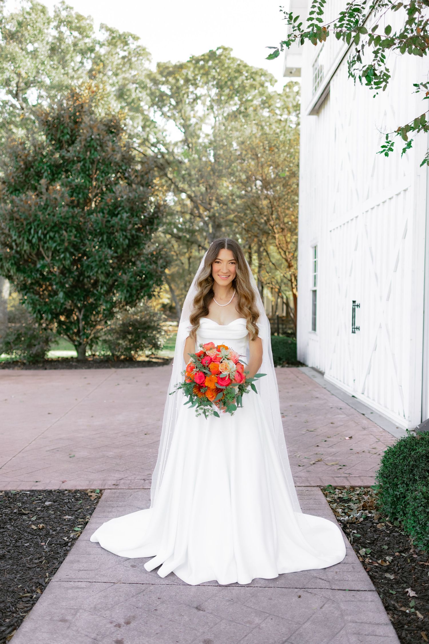 A bride in a flowing white wedding gown holds vibrant orange and red bouquet while standing on a brick pathway.