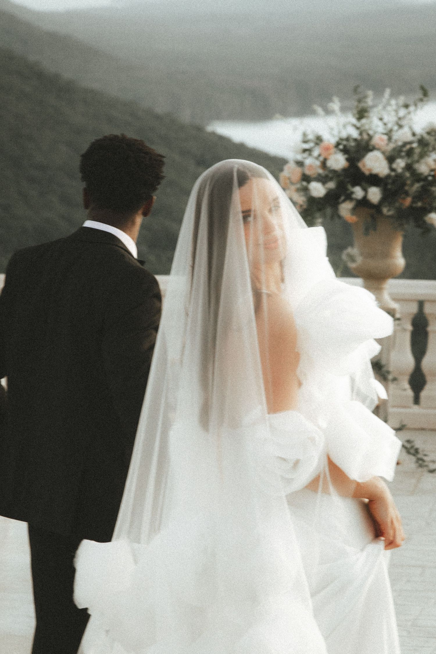 Bride in white gown and veil looks back over shoulder beside groom in black suit on scenic mountain terrace.