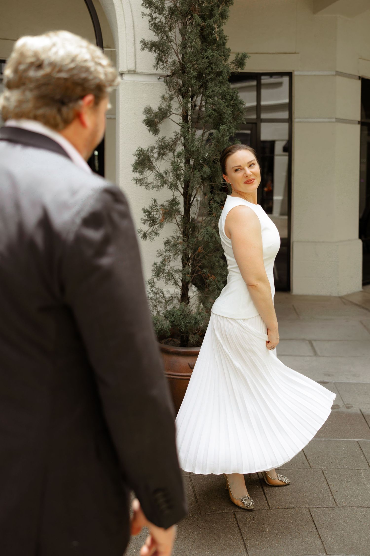 Woman in white pleated dress smiling over shoulder at man in suit on outdoor walkway.