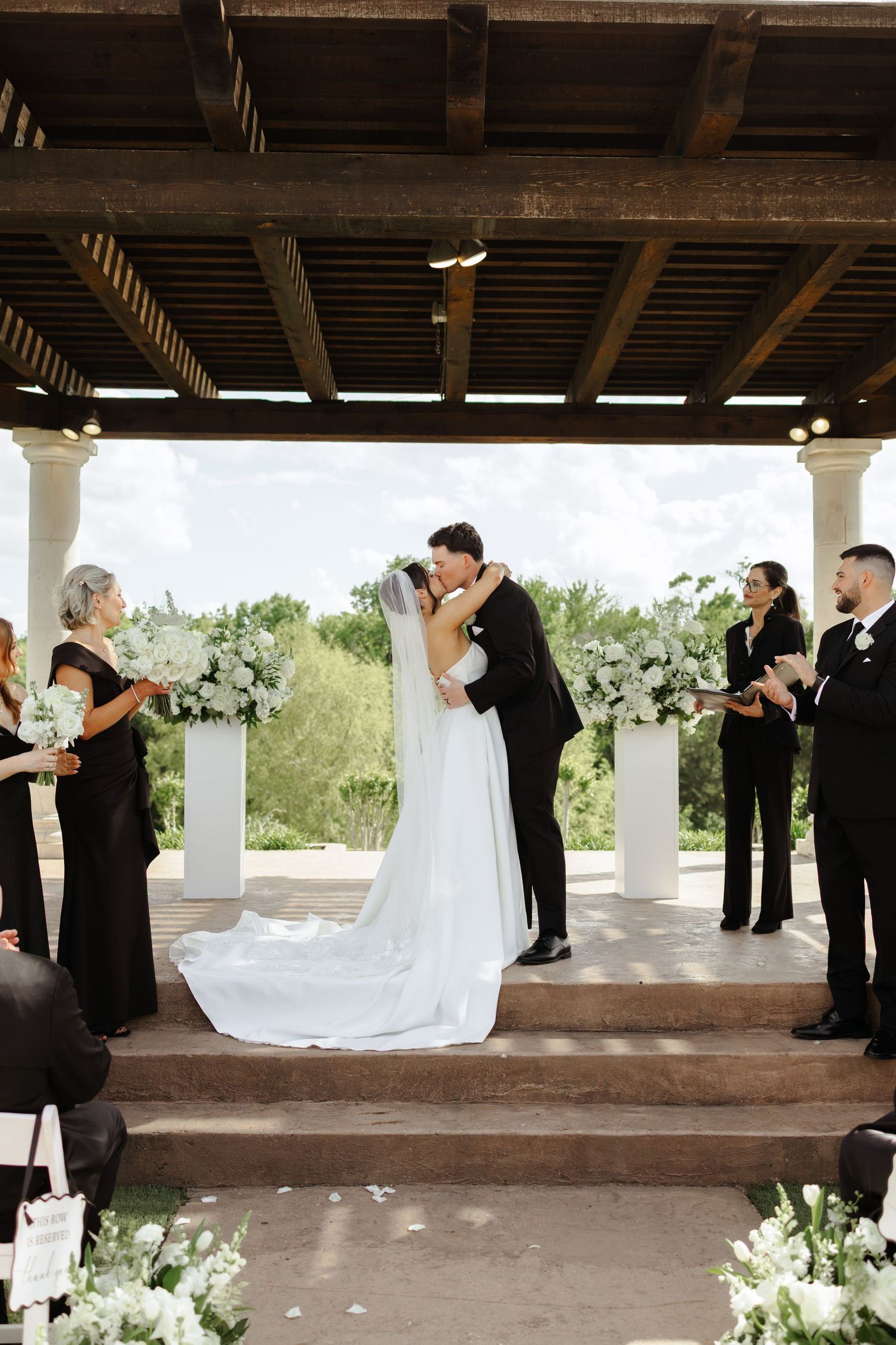Bride and groom share first kiss at outdoor wedding ceremony under pergola with white floral arrangements and wedding party.