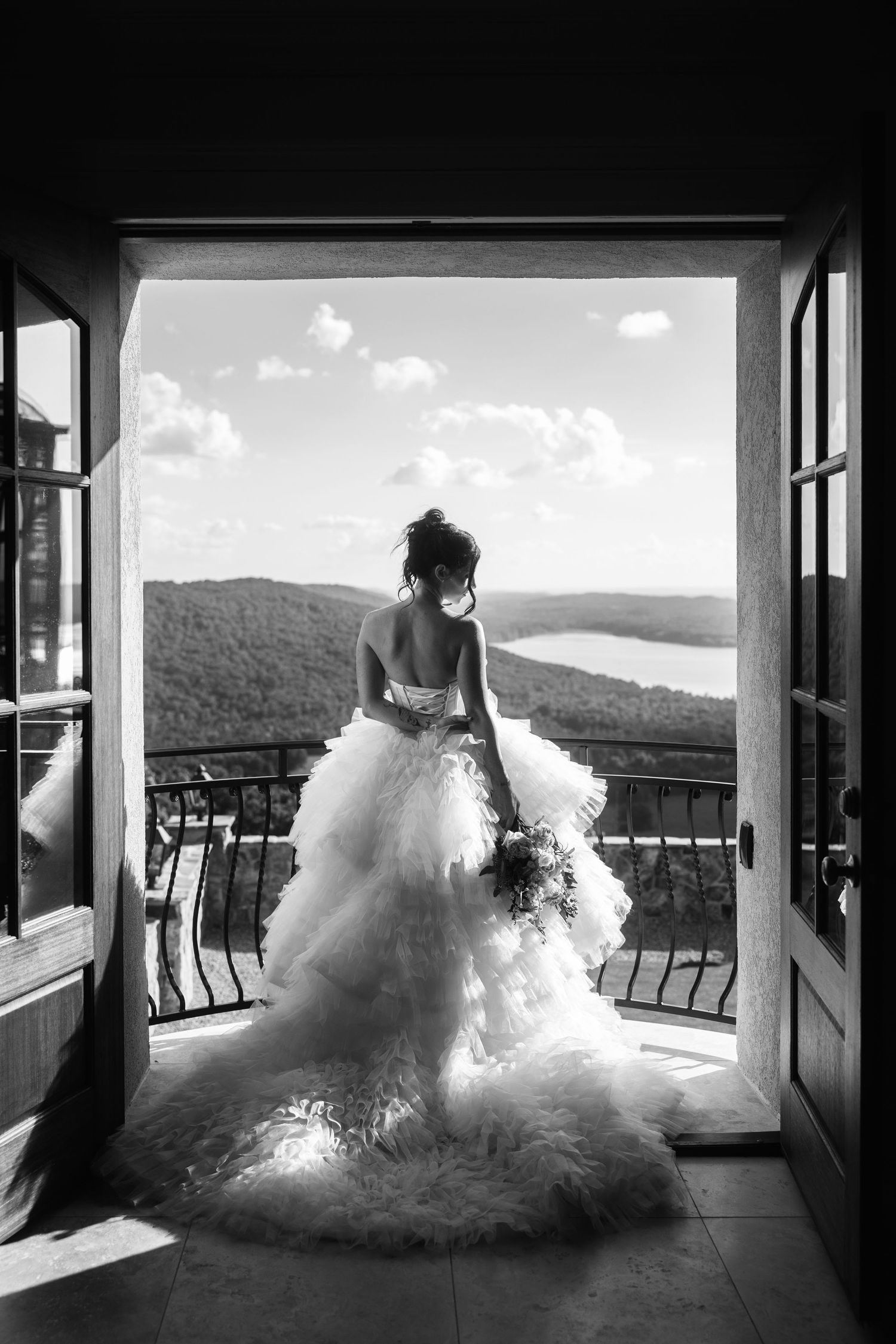 Black and white photo of bride in ruffled ball gown standing in open doorway overlooking lake and mountains.