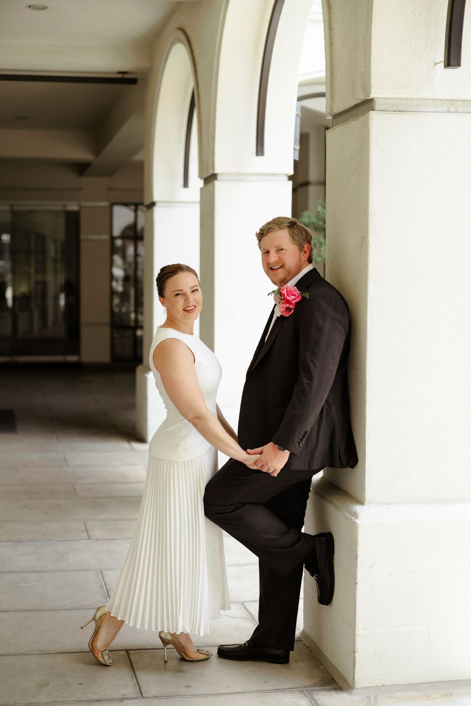 Bride in white pleated dress and groom in black suit with pink boutonniere posing by white columns.